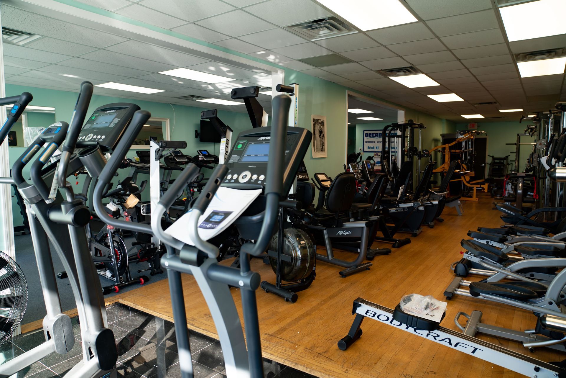 A gym filled with various exercise machines, including elliptical trainers and stationary bikes, on a wood floor.