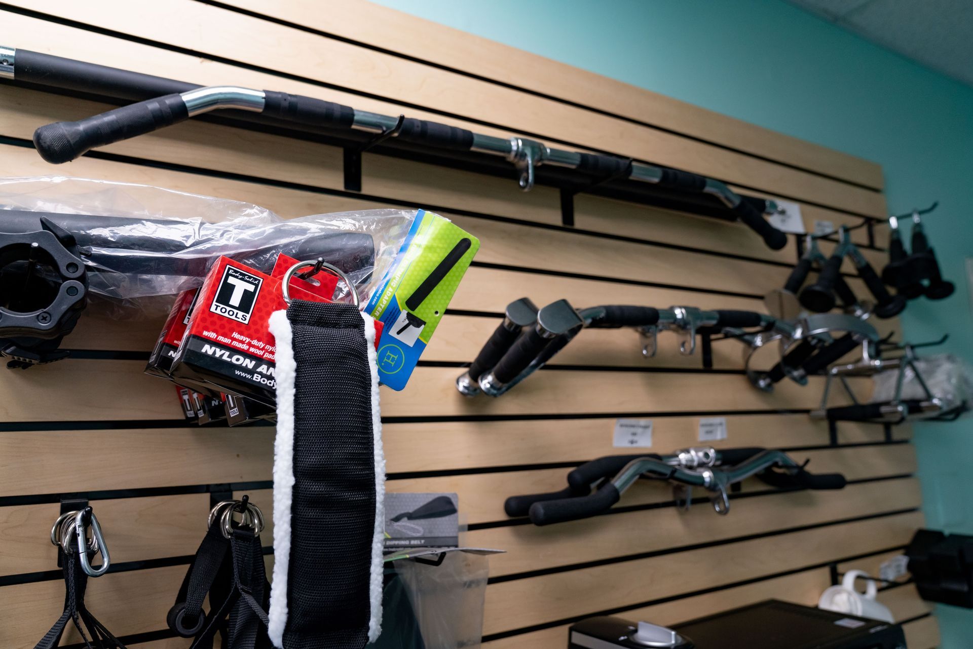 A display wall in a gym featuring various metal cable machine attachments and handles hanging on horizontal slats.