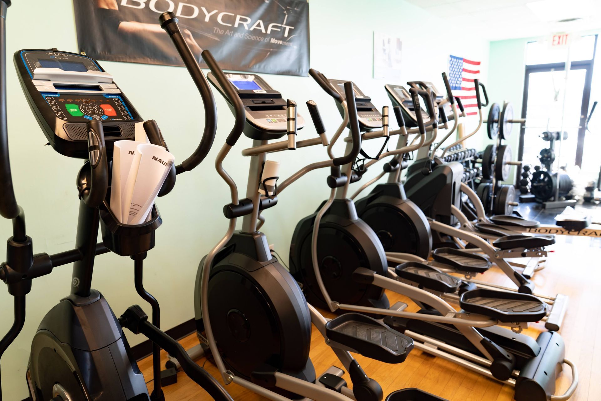 A row of gray elliptical exercise machines lined up in a brightly lit gym with a Bodycraft sign on the wall.
