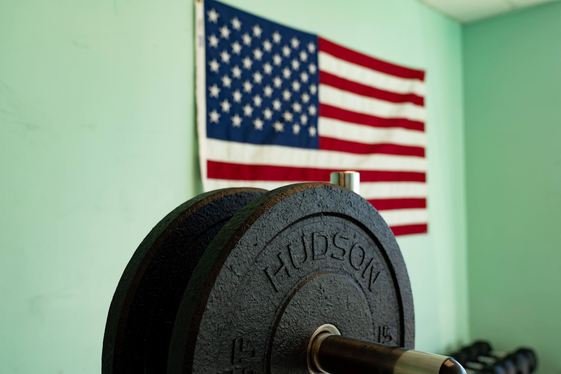 Hudson weight plates on a barbell in a gym with an American flag hanging on a light green wall in the background.