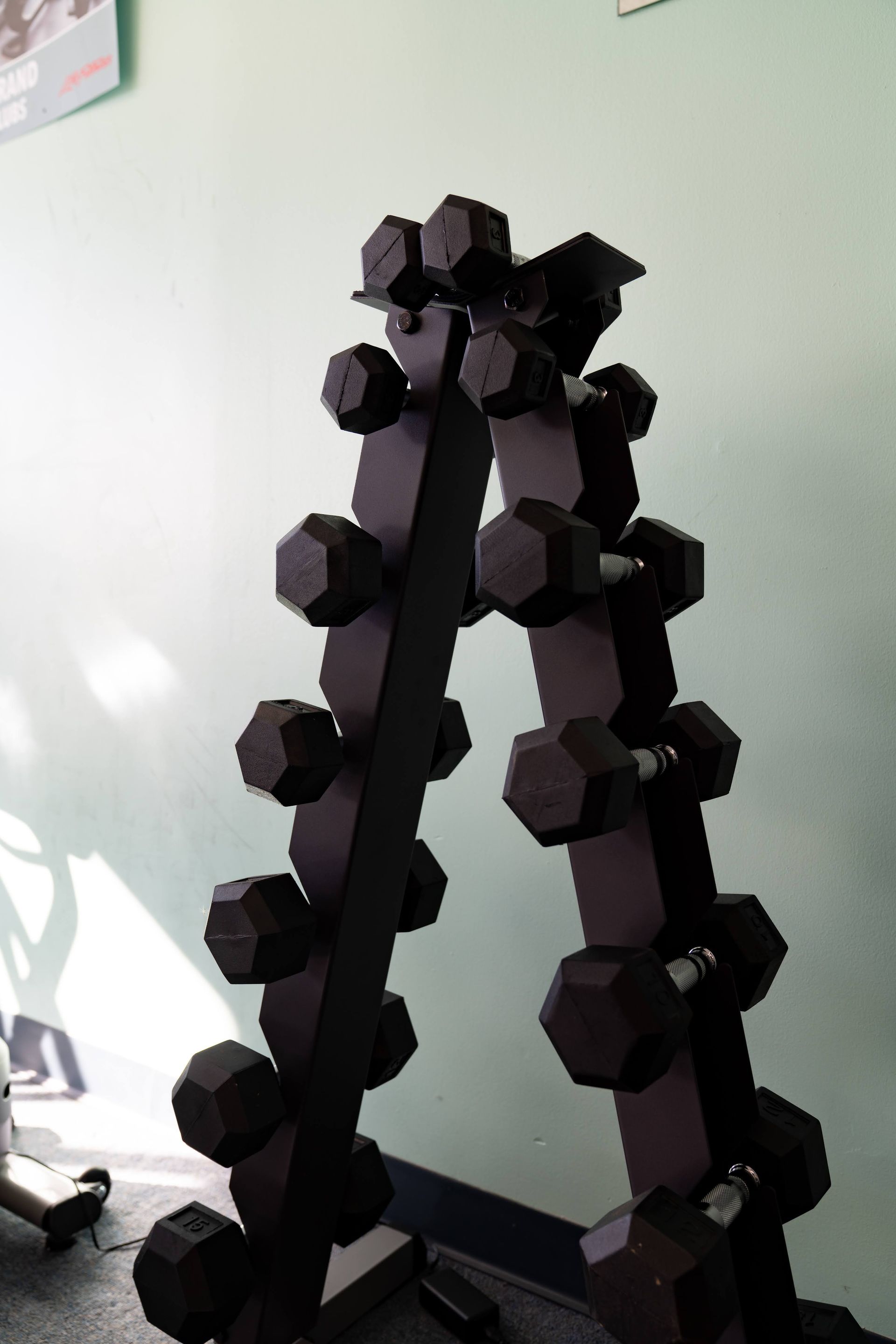 A black A-frame dumbbell rack stands against a light green wall, holding several pairs of black hexagonal dumbbells.