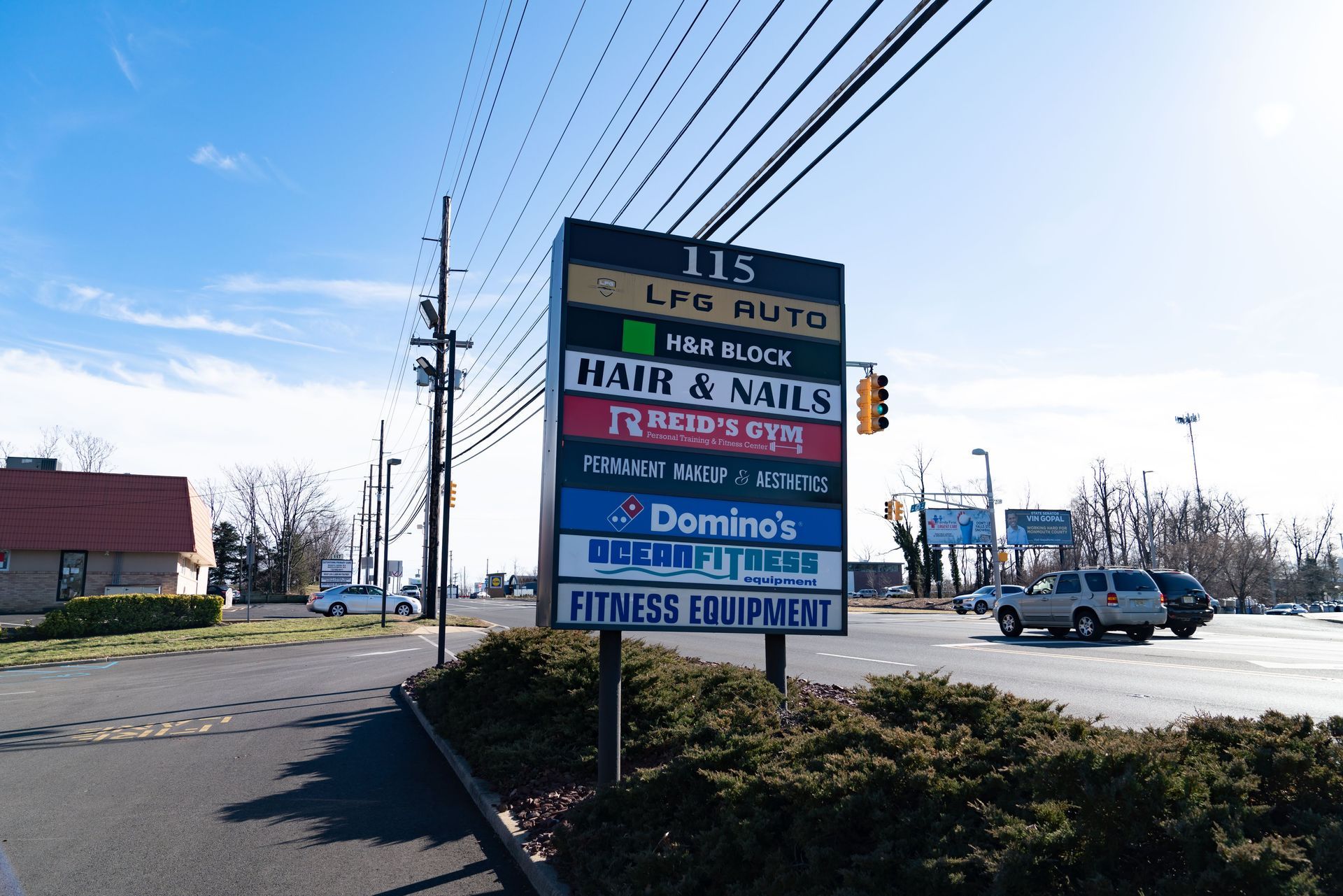 A multi-tenant sign at 115 listing businesses including Domino's, a hair salon, and fitness equipment, under a blue sky.