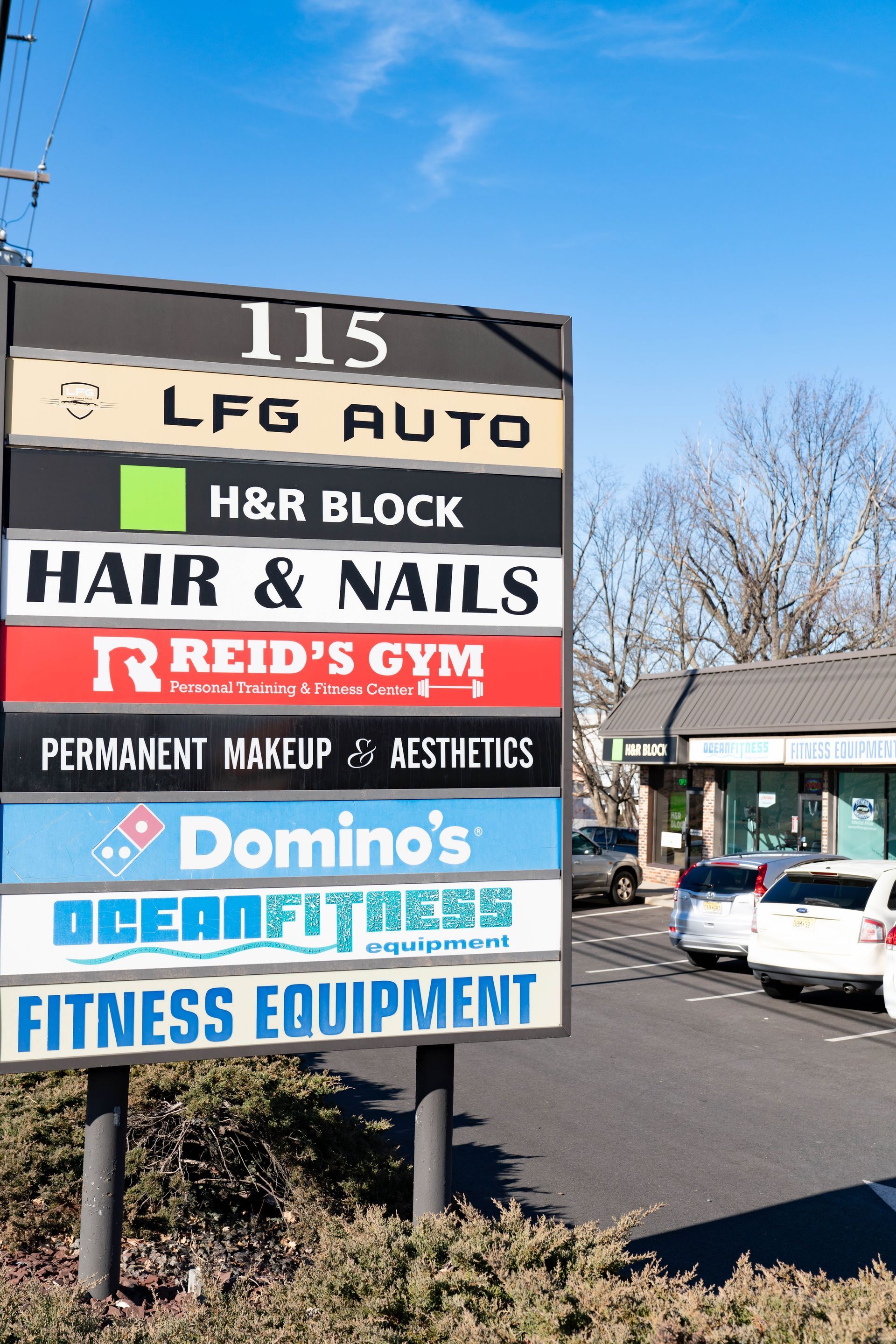 A sign listing various businesses at 115, including LFG Auto, H&R Block, Hair & Nails, Reid's Gym, and Domino's.