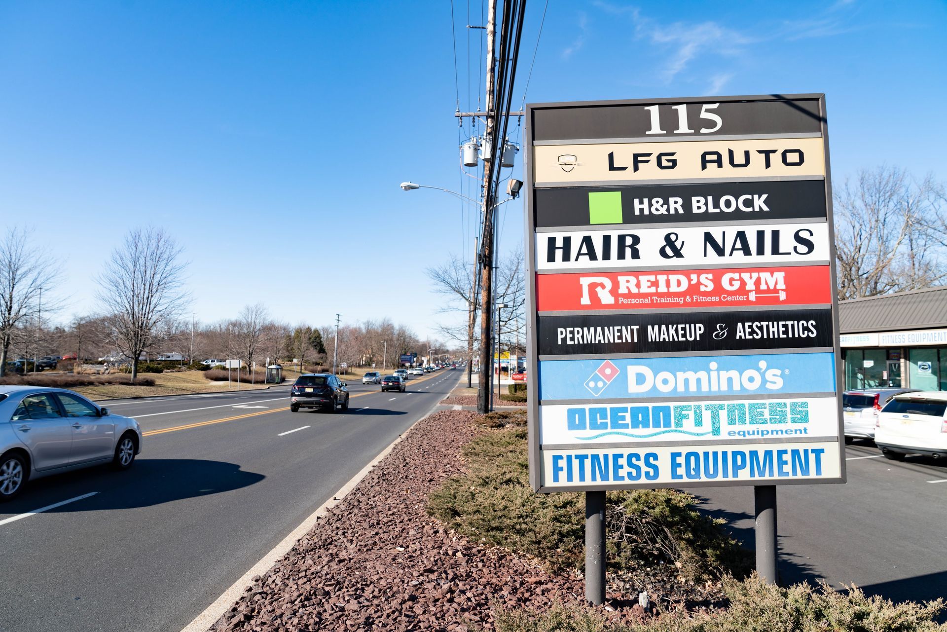 A tall commercial directory sign at 115 listing several businesses along a sunny roadside with cars passing by.