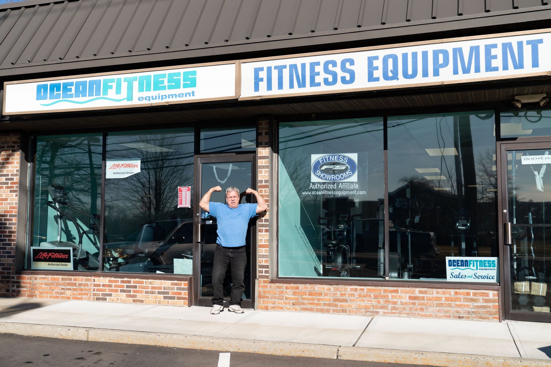 A person standing in the doorway of Ocean Fitness, flexing their arms in front of the store with fitness equipment inside.