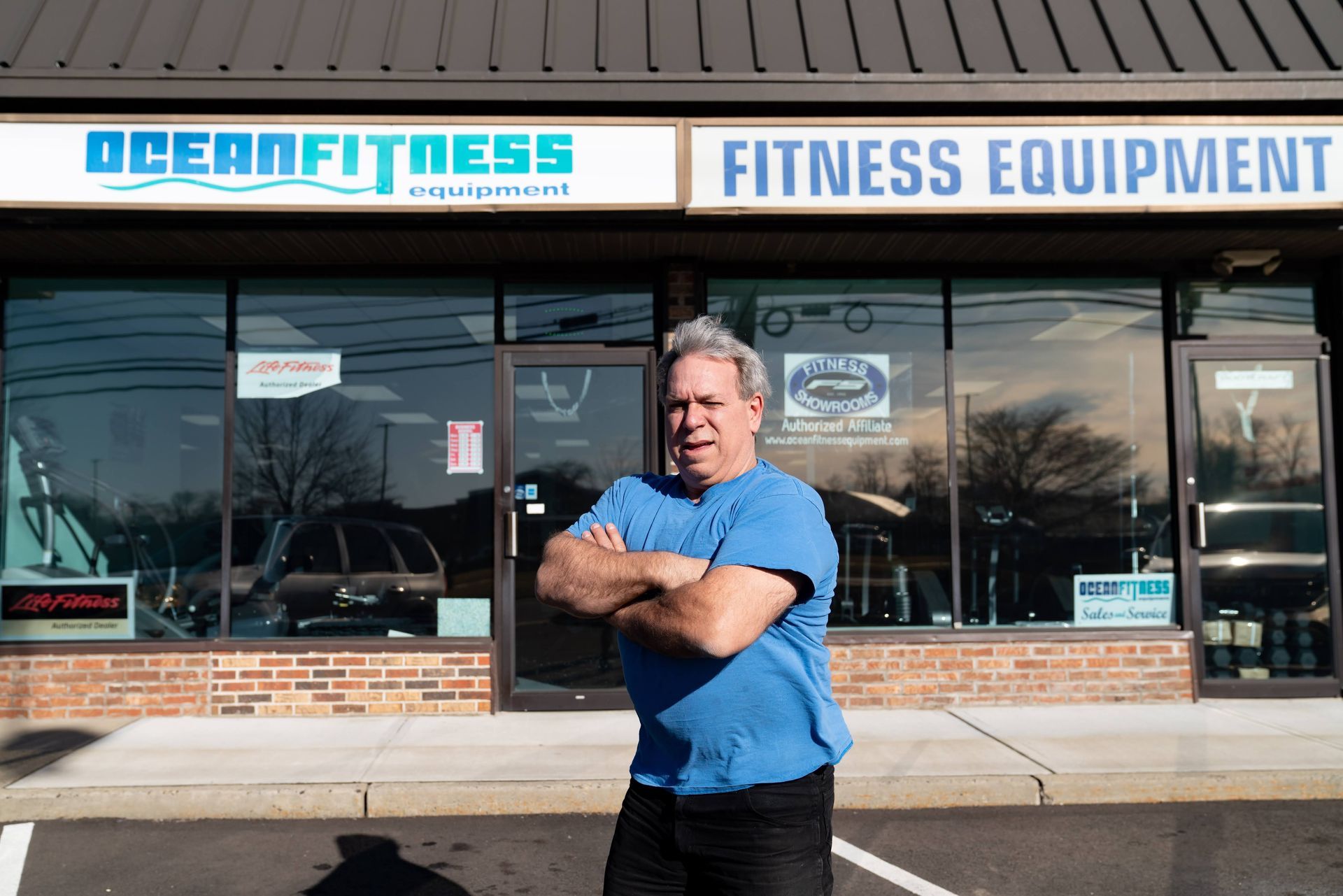 A person with arms crossed stands in front of a store named Ocean Fitness Equipment with large glass windows.