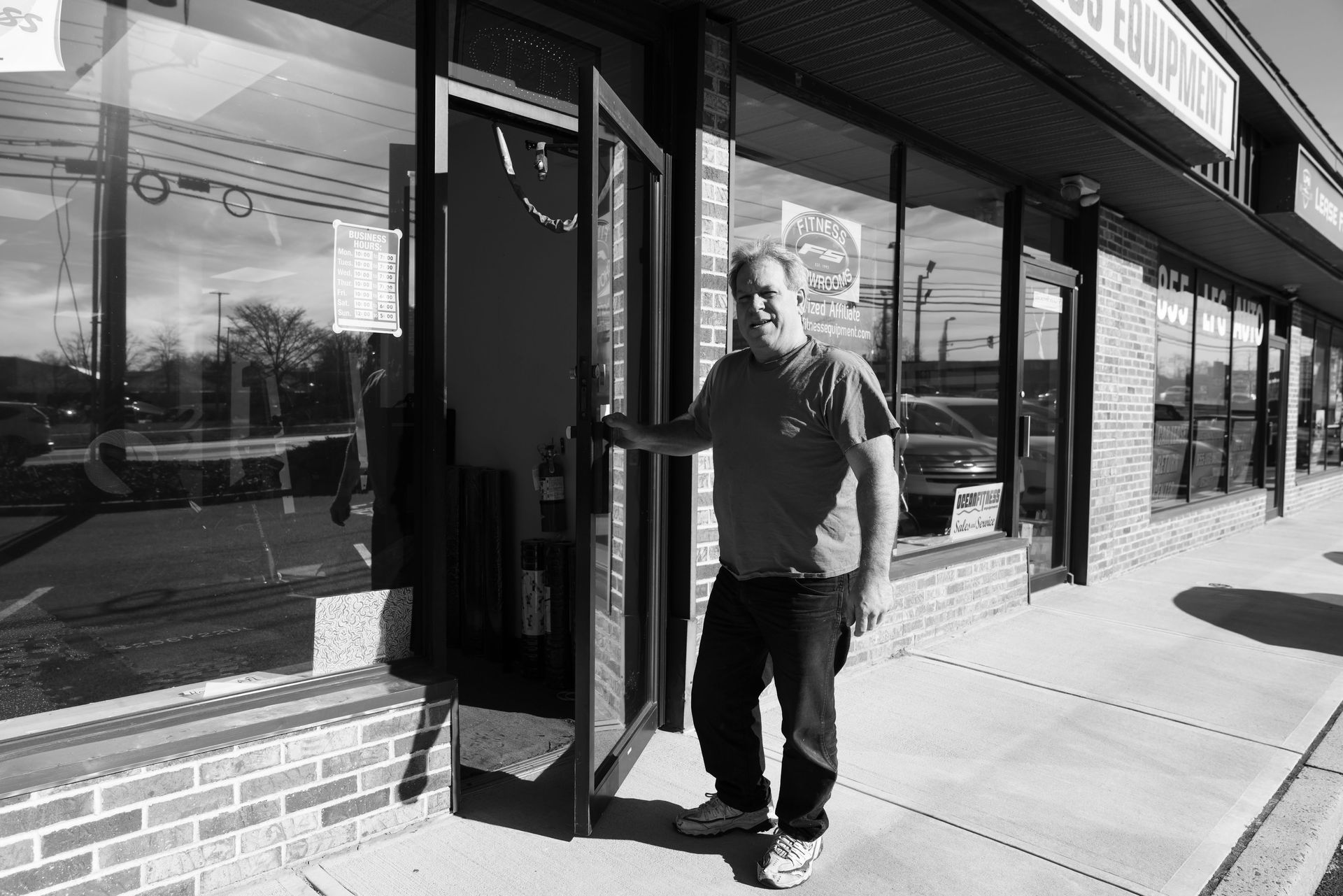 A man stands in an open doorway of a storefront in a strip mall, smiling and holding the door open.