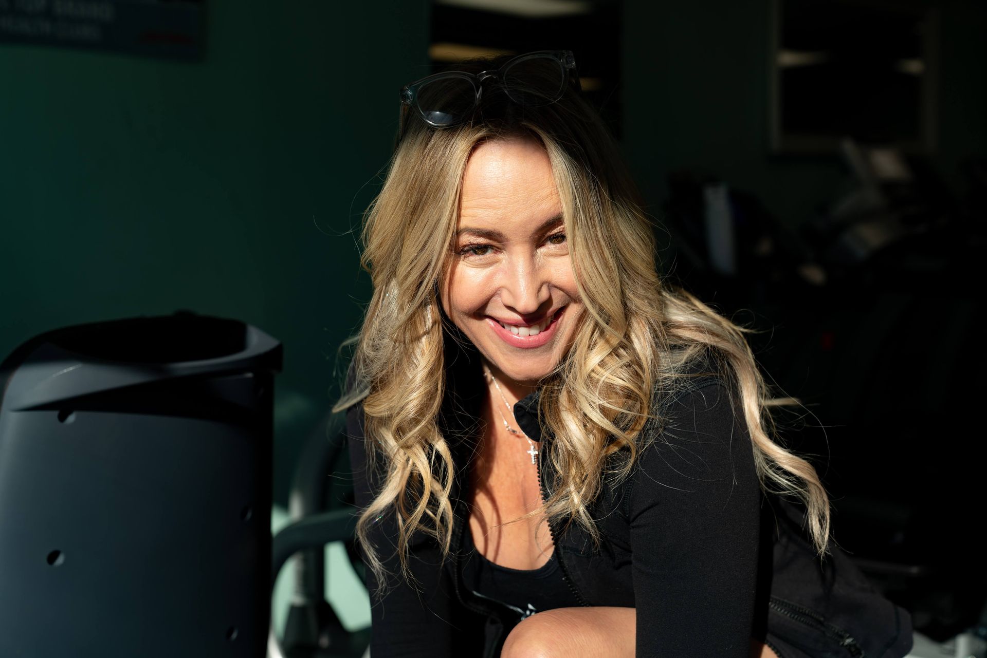 A smiling blonde person with wavy hair, wearing a black zip-up jacket, looking at the camera in a dimly lit room.