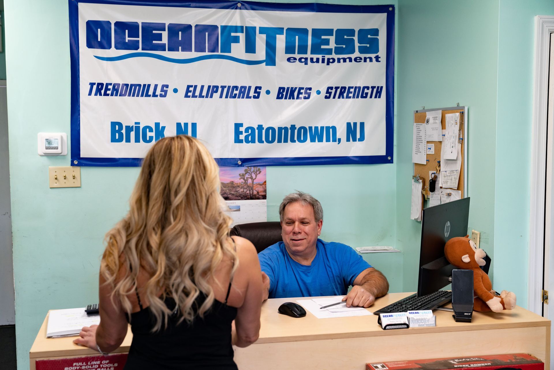 A person stands at a desk in an Ocean Fitness Equipment office, speaking with a staff member in a blue shirt.