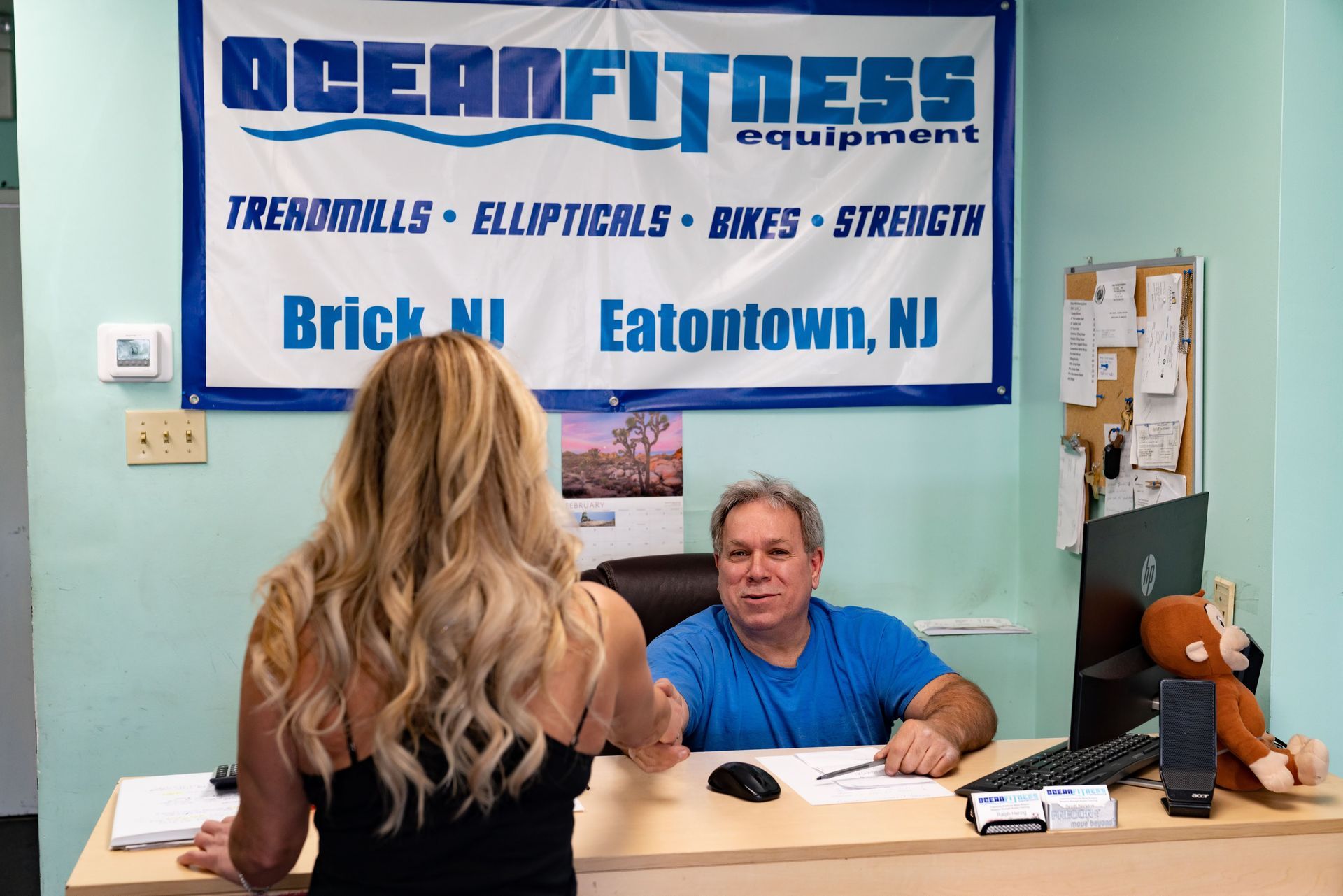 A person shaking hands with a man sitting at a desk under an Ocean Fitness Equipment sign in an office setting.