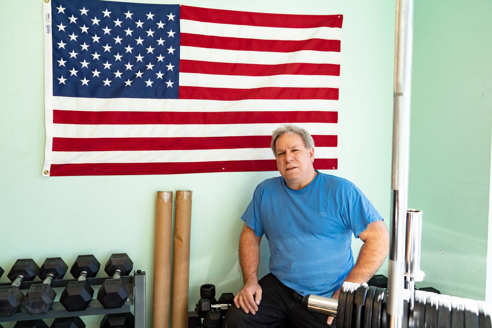 A person wearing a blue t-shirt sits in a gym in front of an American flag hung on a light green wall.