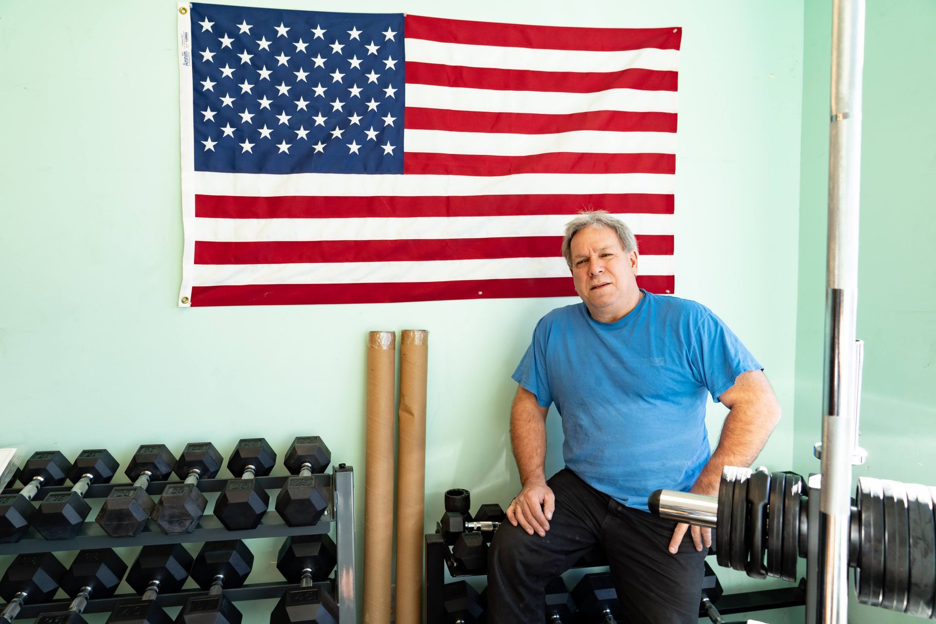 A person in a blue t-shirt sits in a home gym in front of an American flag, with dumbbells on a rack in the foreground.