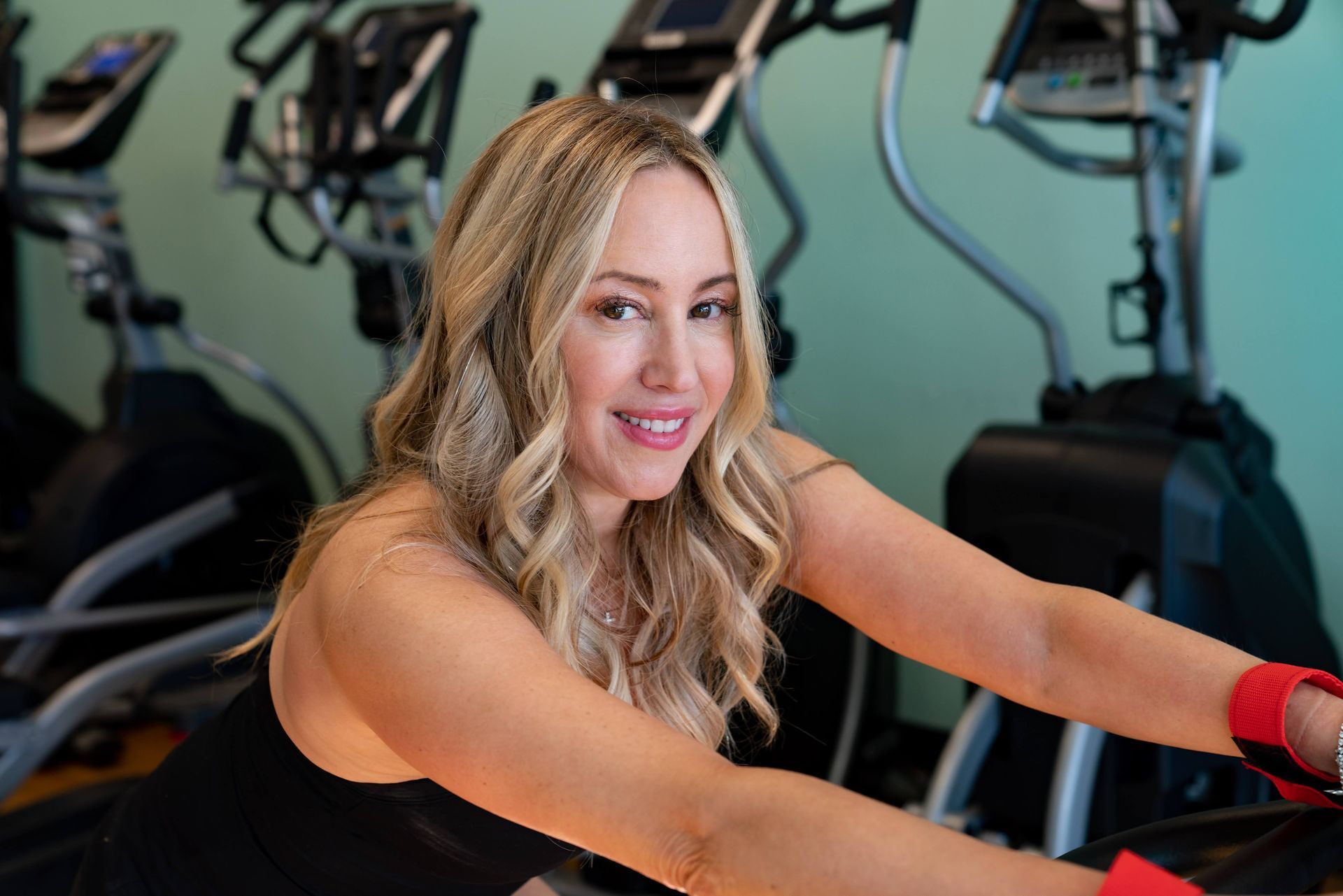 A person with blonde, wavy hair smiles while exercising on a stationary machine in a gym setting.