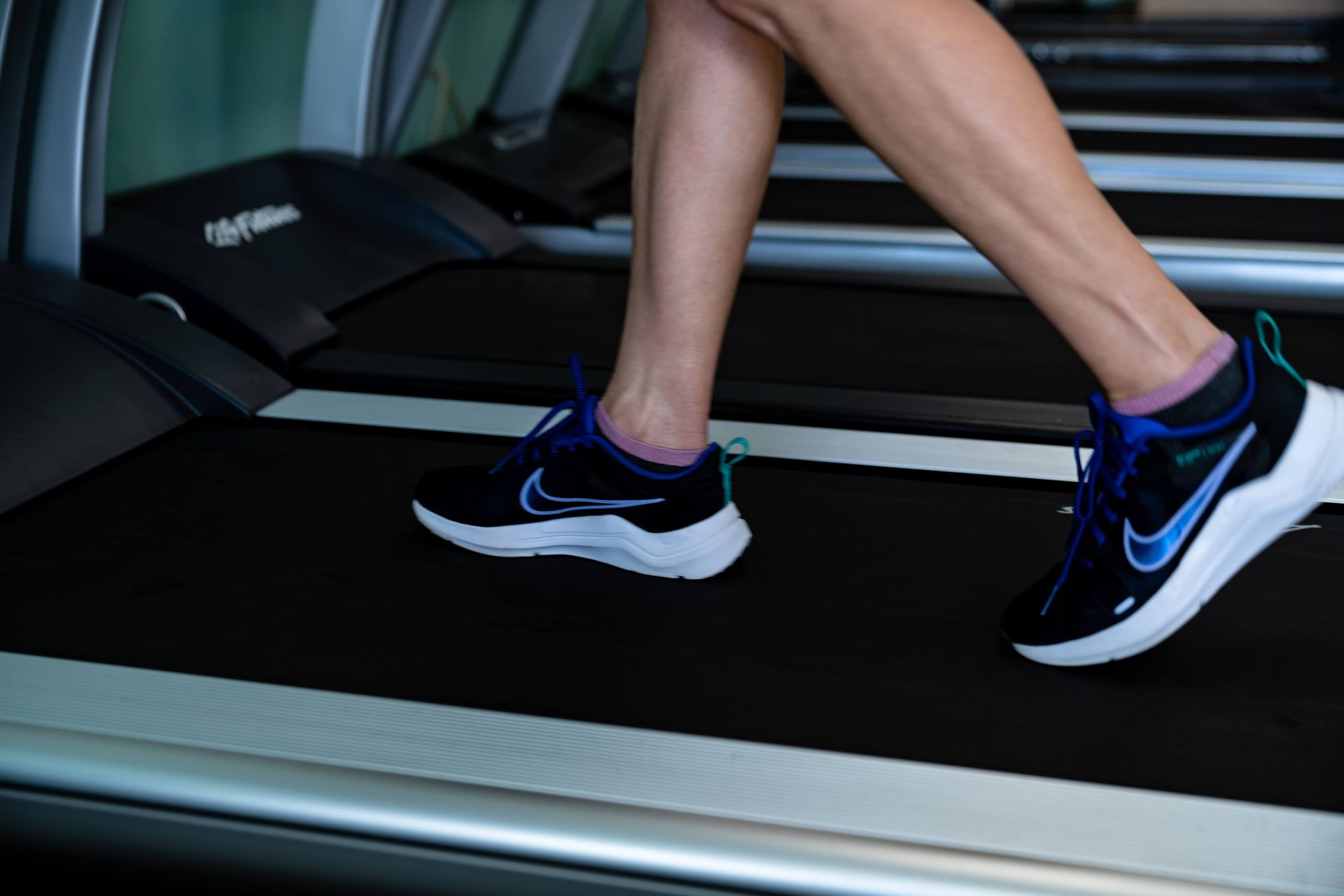 A person running on a gym treadmill, wearing black Nike sneakers with blue accents and light pink ankle socks.