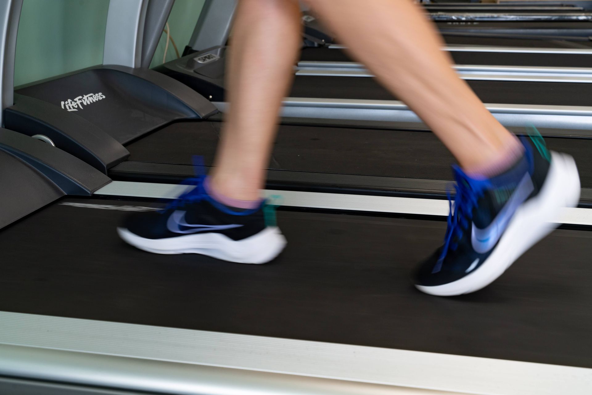 A person wearing blue and black sneakers runs on a treadmill in a gym setting.