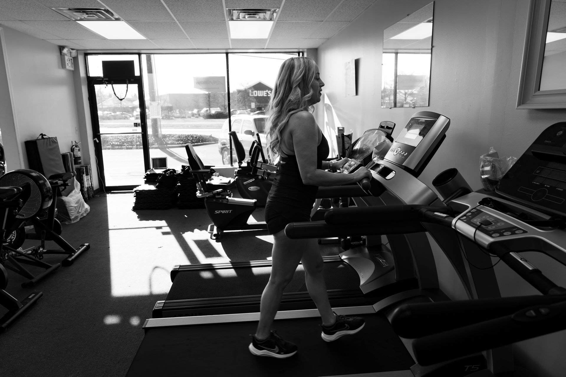 A person walks on a treadmill in a gym with large windows in the background.