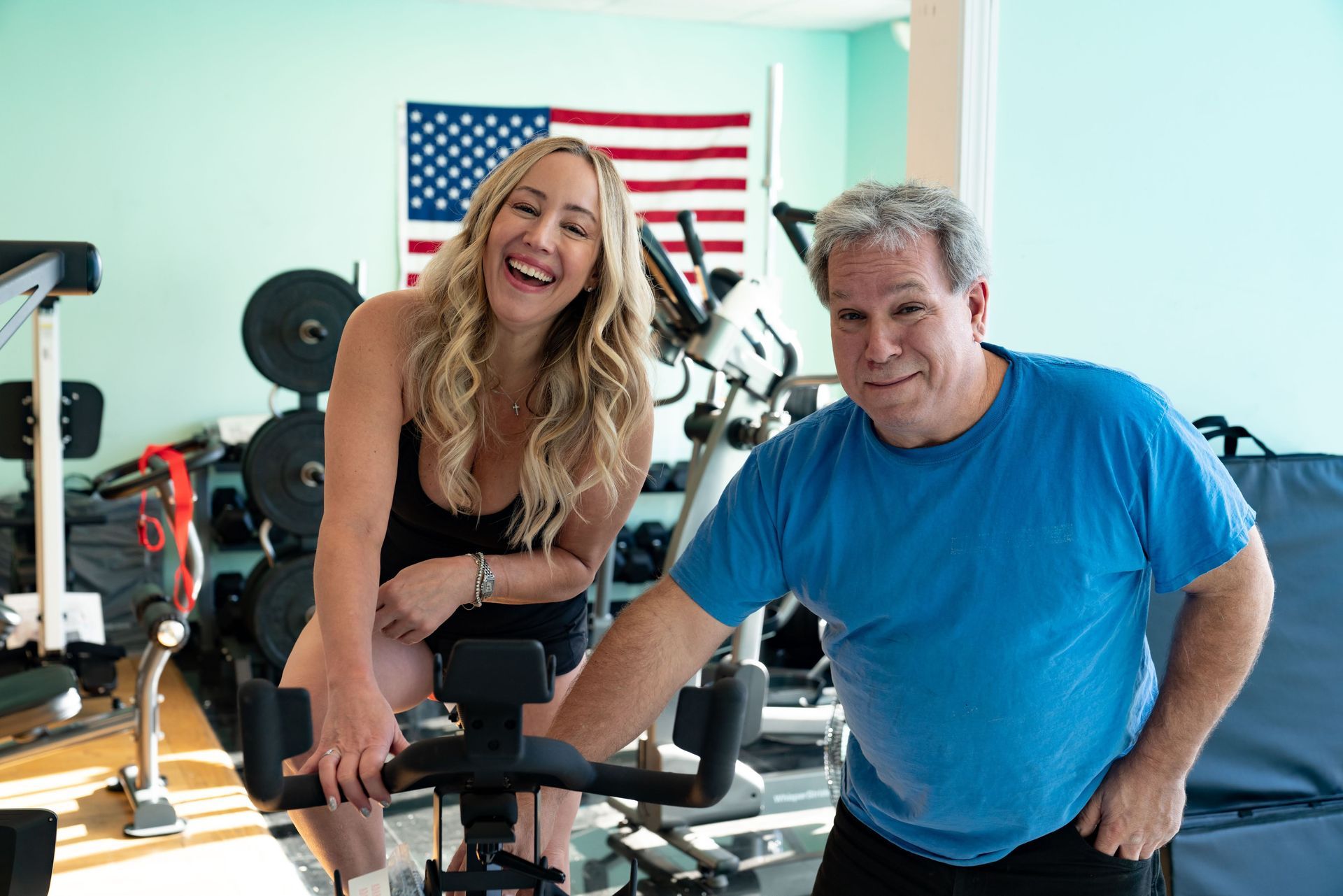 A smiling woman and a man in a blue shirt stand next to an indoor exercise bike in a gym with an American flag background.