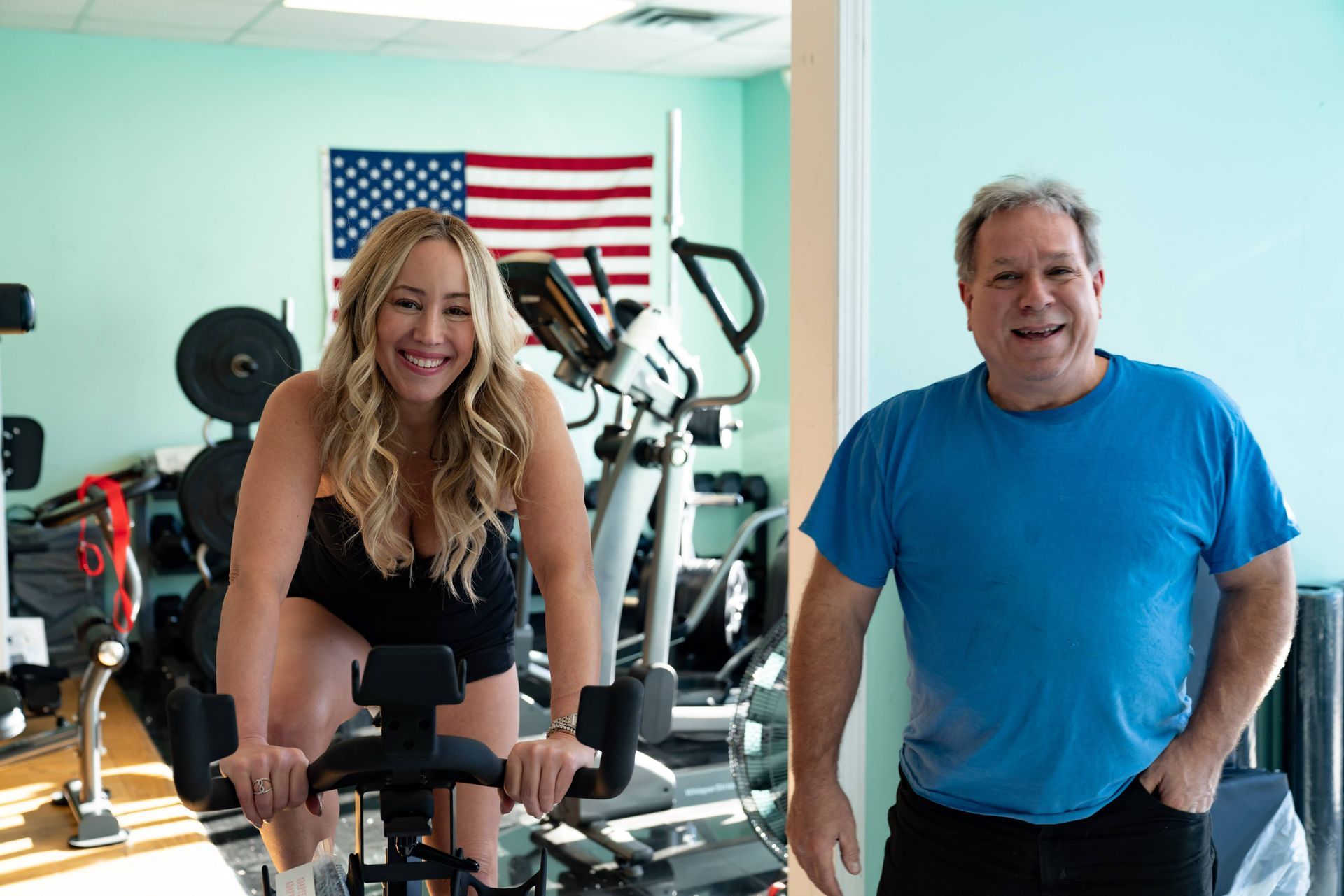 A smiling person rides a stationary bike in a gym, with another person standing nearby wearing a blue shirt.