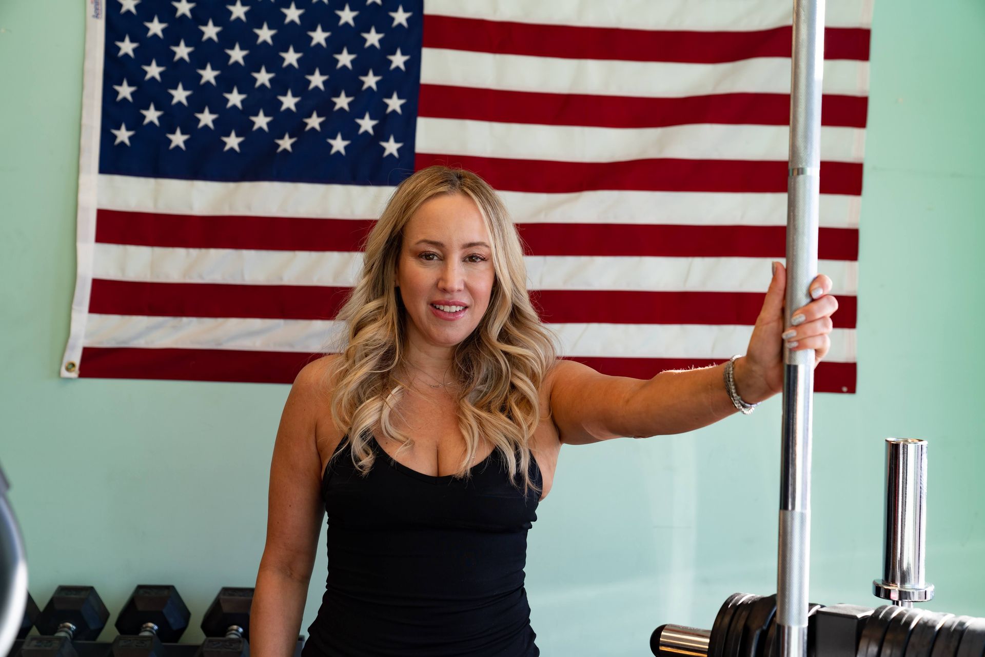 A smiling person in a black tank top stands in a gym in front of an American flag, holding a vertical barbell.