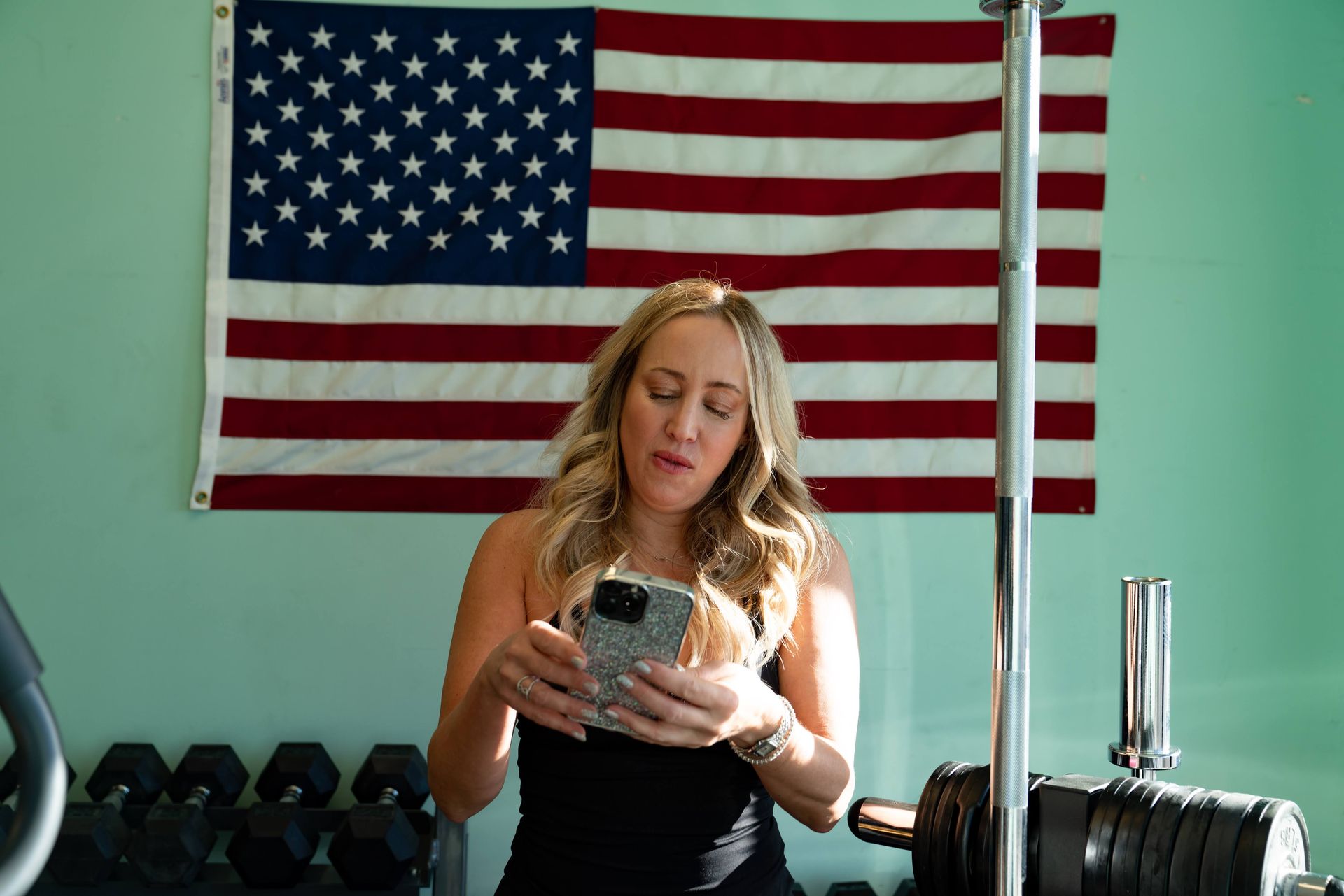 A person holds a phone in a home gym with an American flag on the wall and weightlifting equipment in the background.