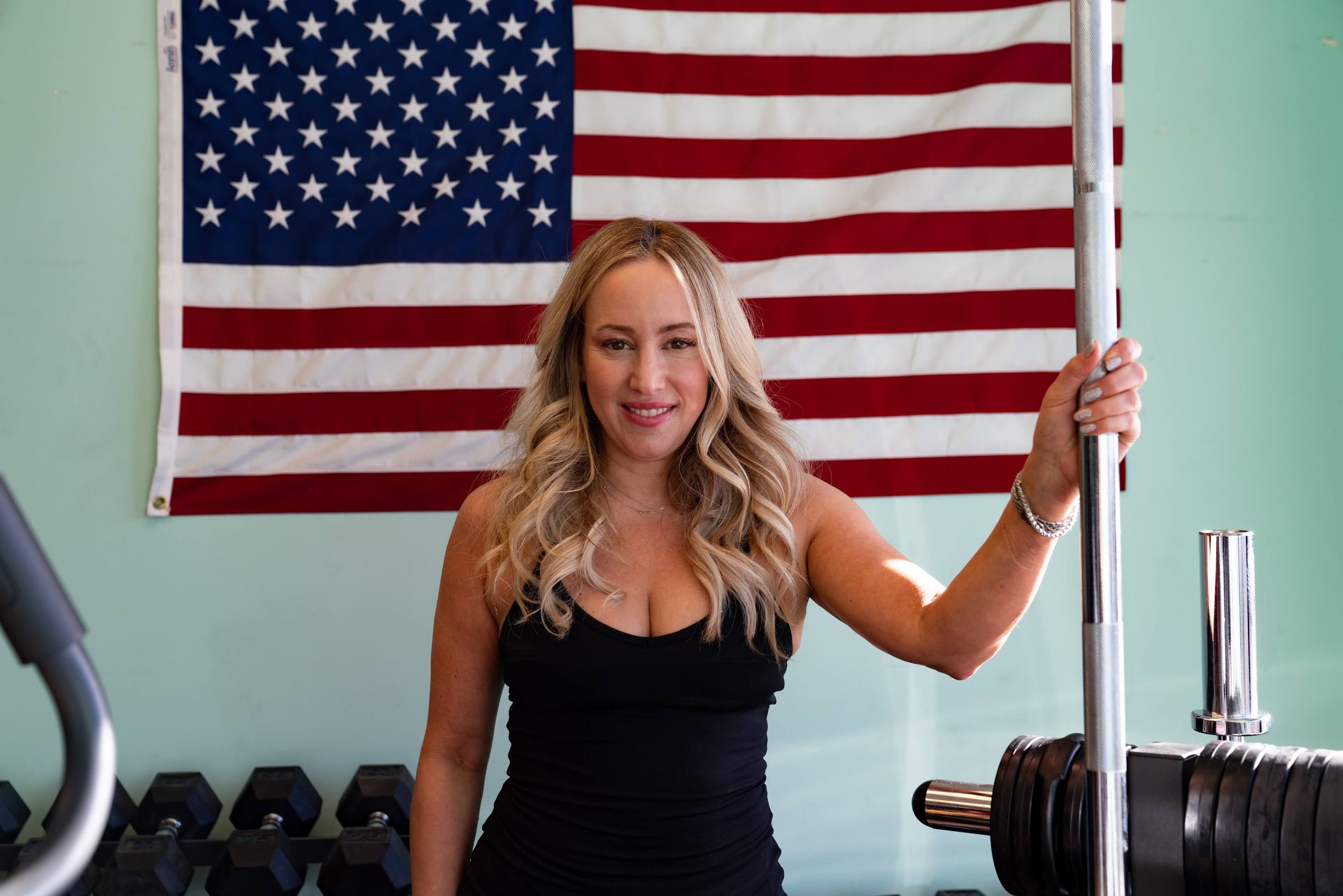 A smiling person in a black tank top stands in a gym, holding a vertical barbell in front of an American flag.