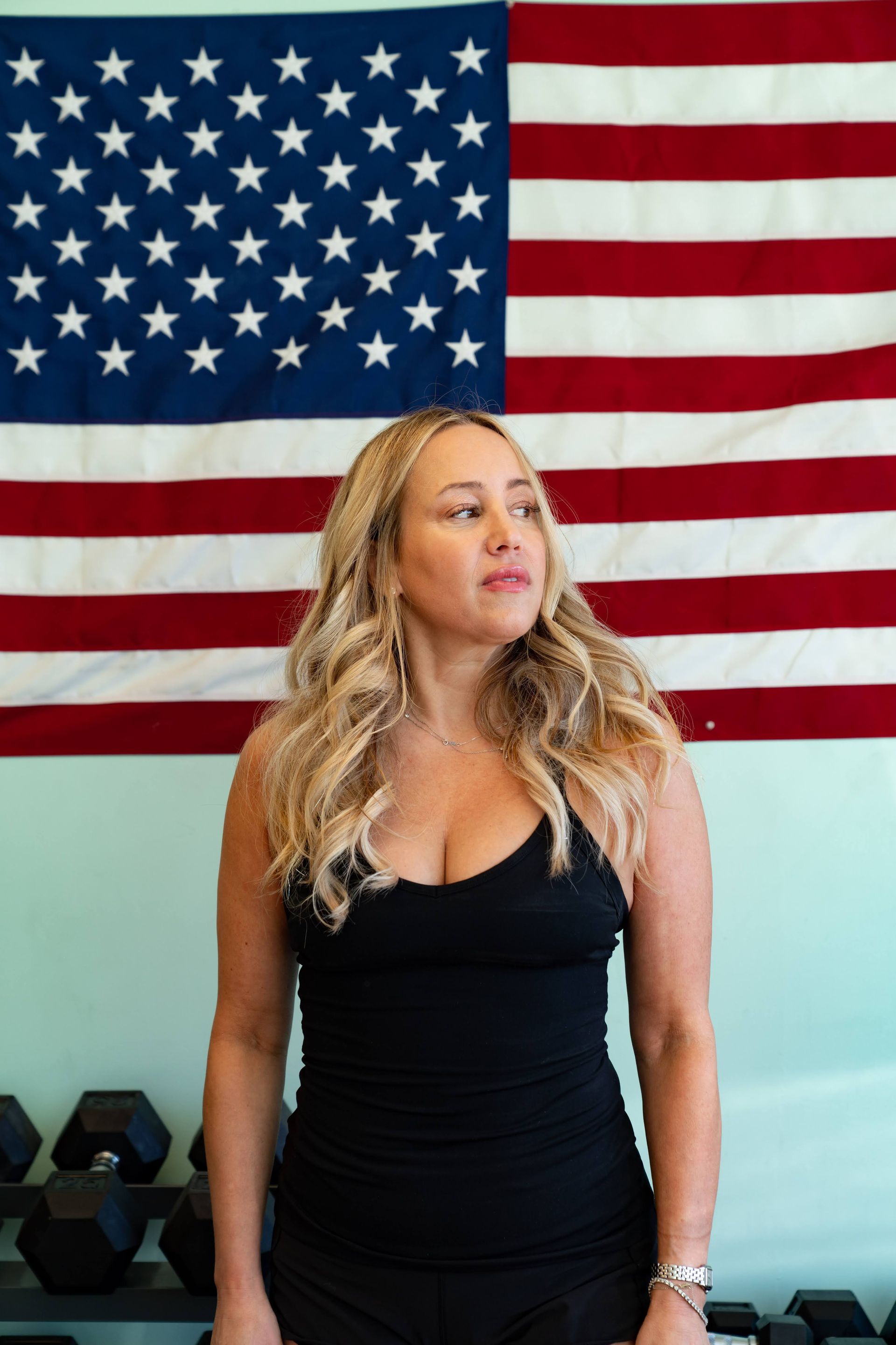 A blonde person in a black tank top stands before an American flag, looking to the side with dumbbells in the foreground.