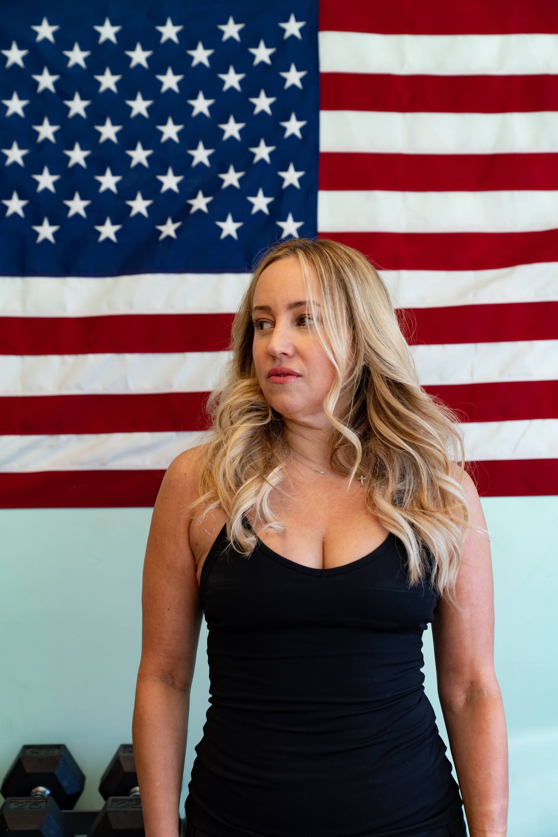 A person with shoulder-length blonde hair in a black tank top stands before a large American flag.