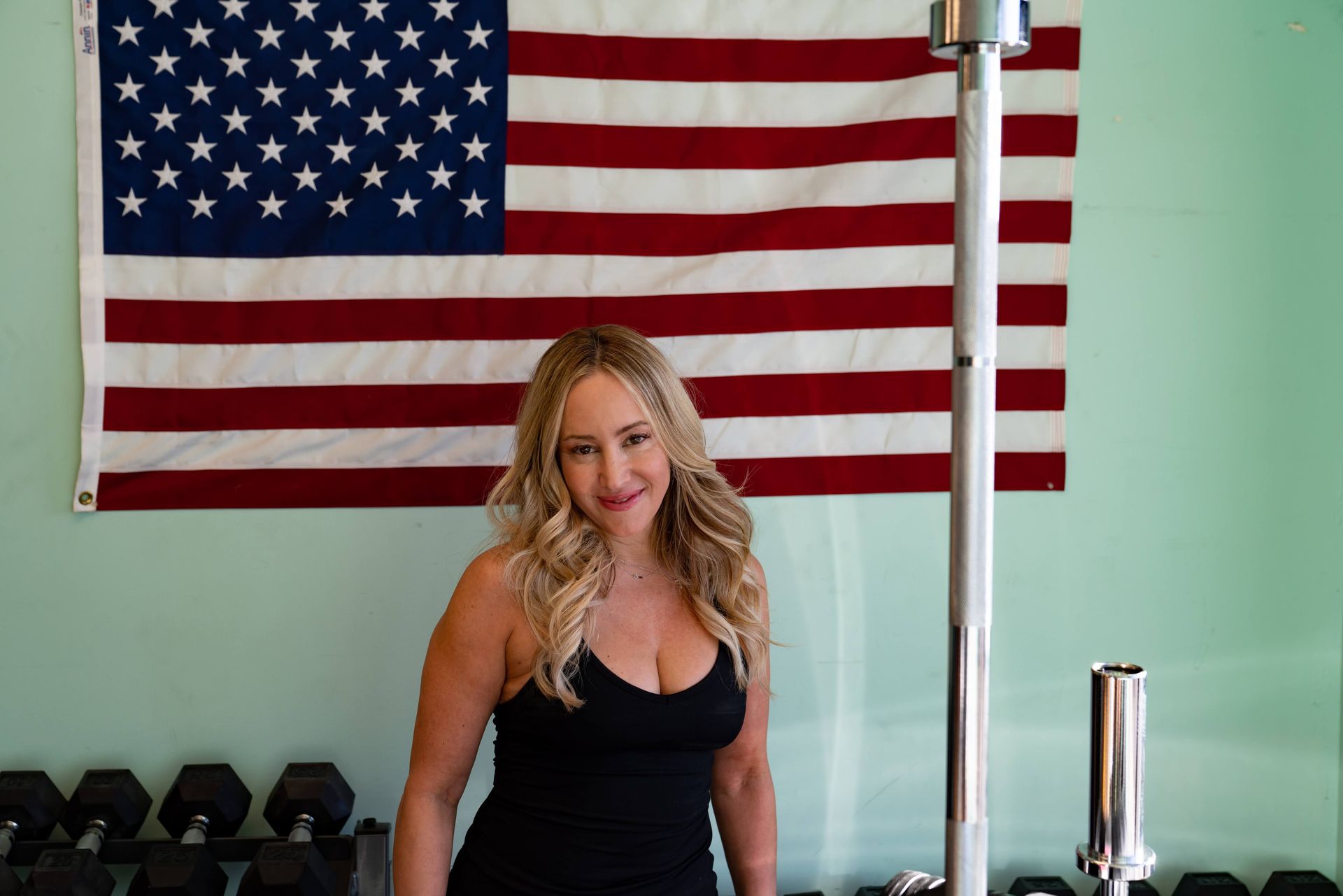 A smiling woman stands in front of a wall-mounted American flag in a gym, with barbells and weights visible nearby.