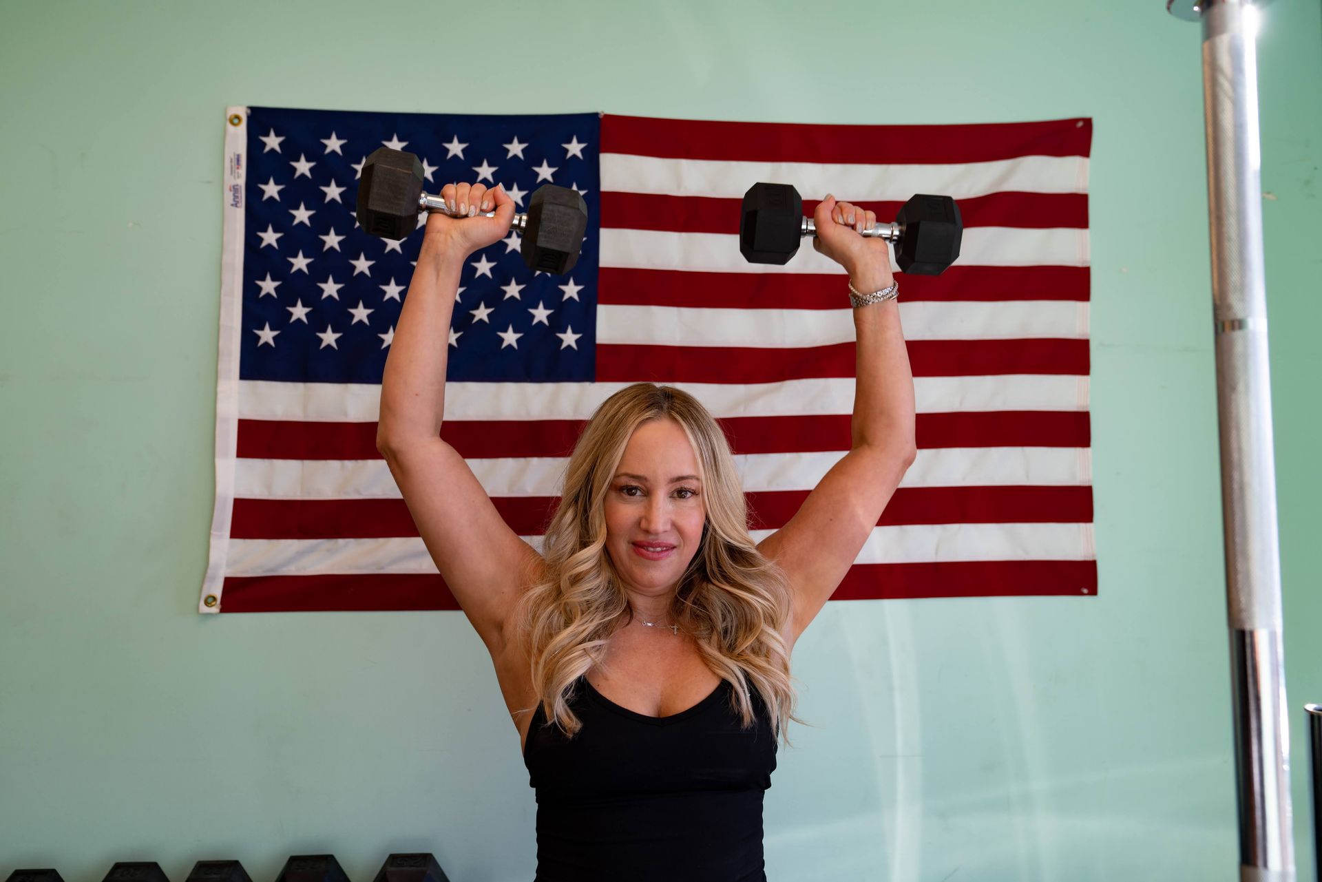 A person standing in front of an American flag, lifting two black dumbbells overhead in a shoulder press exercise.