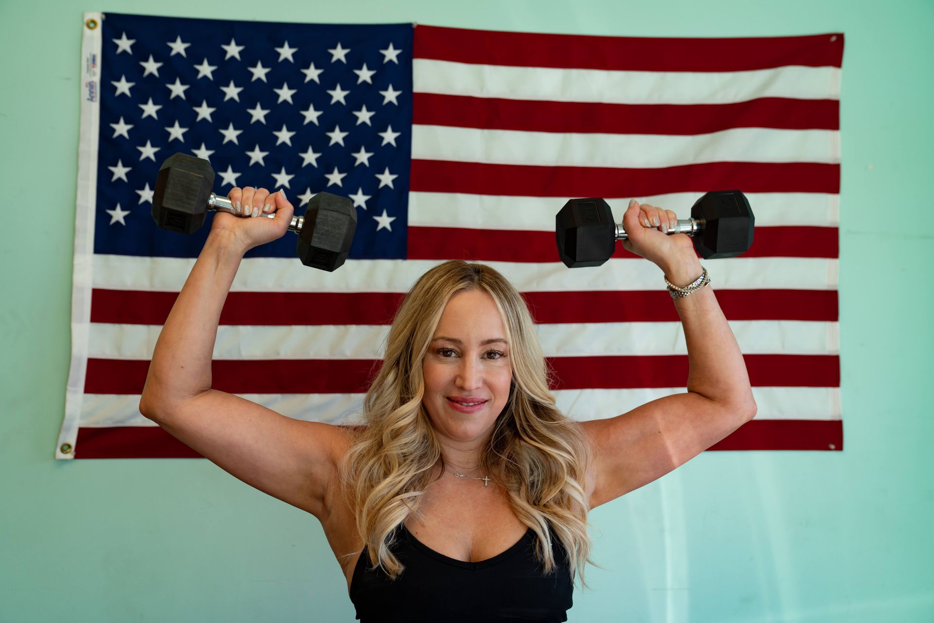 A person smiling while performing an overhead dumbbell press in front of an American flag hanging on a light blue wall.