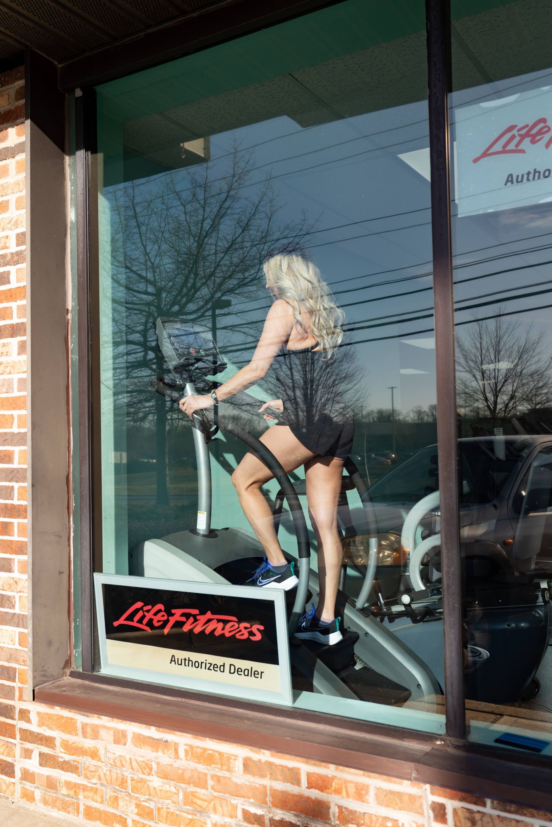 A person in a black outfit exercising on a Life Fitness elliptical machine visible through a store window.