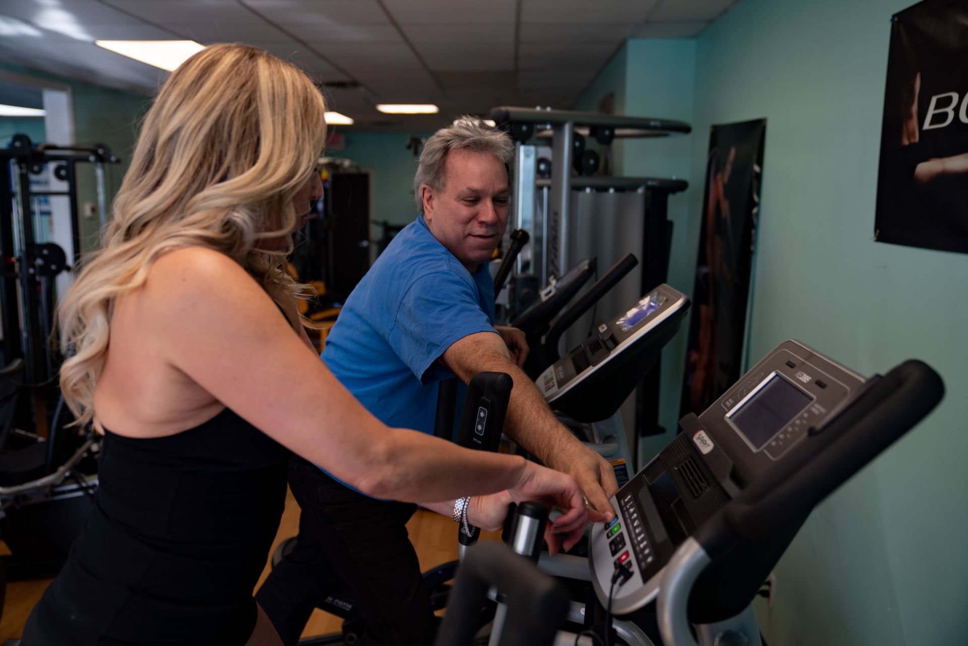 A fitness trainer in a blue shirt assists an individual with using an elliptical machine in a gym.