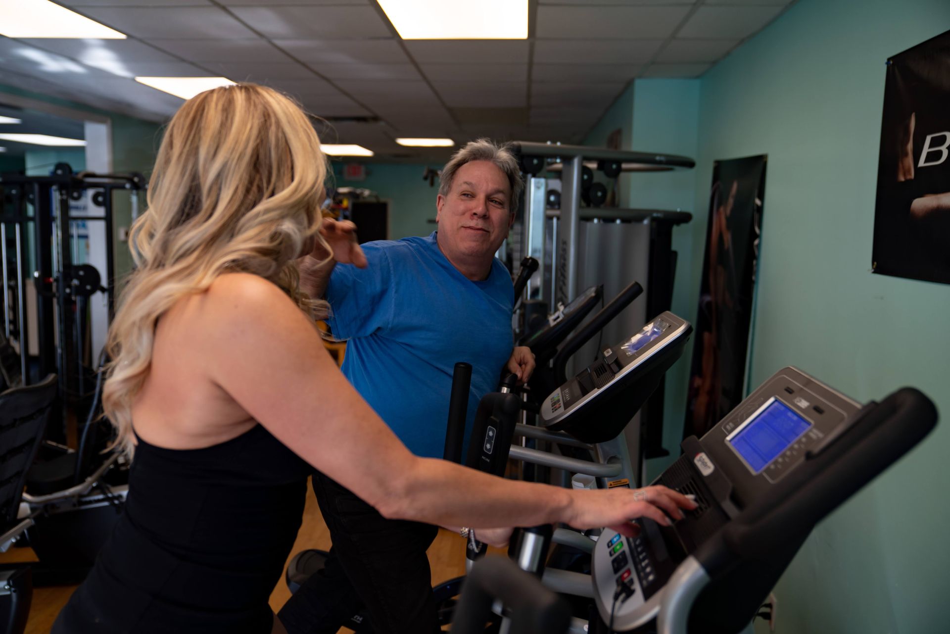 A woman in a black tank top interacts with the screen of an elliptical machine while a man in a blue shirt watches.