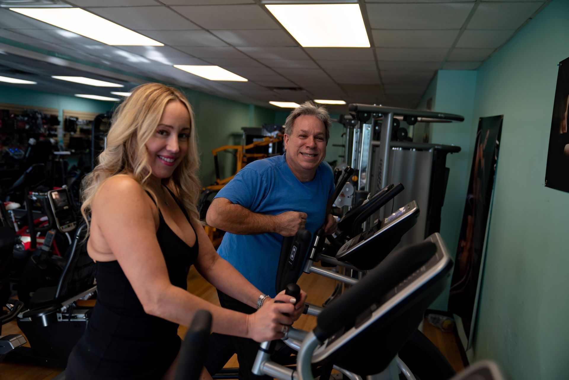 Two people smiling while exercising on elliptical machines in a brightly lit gym.