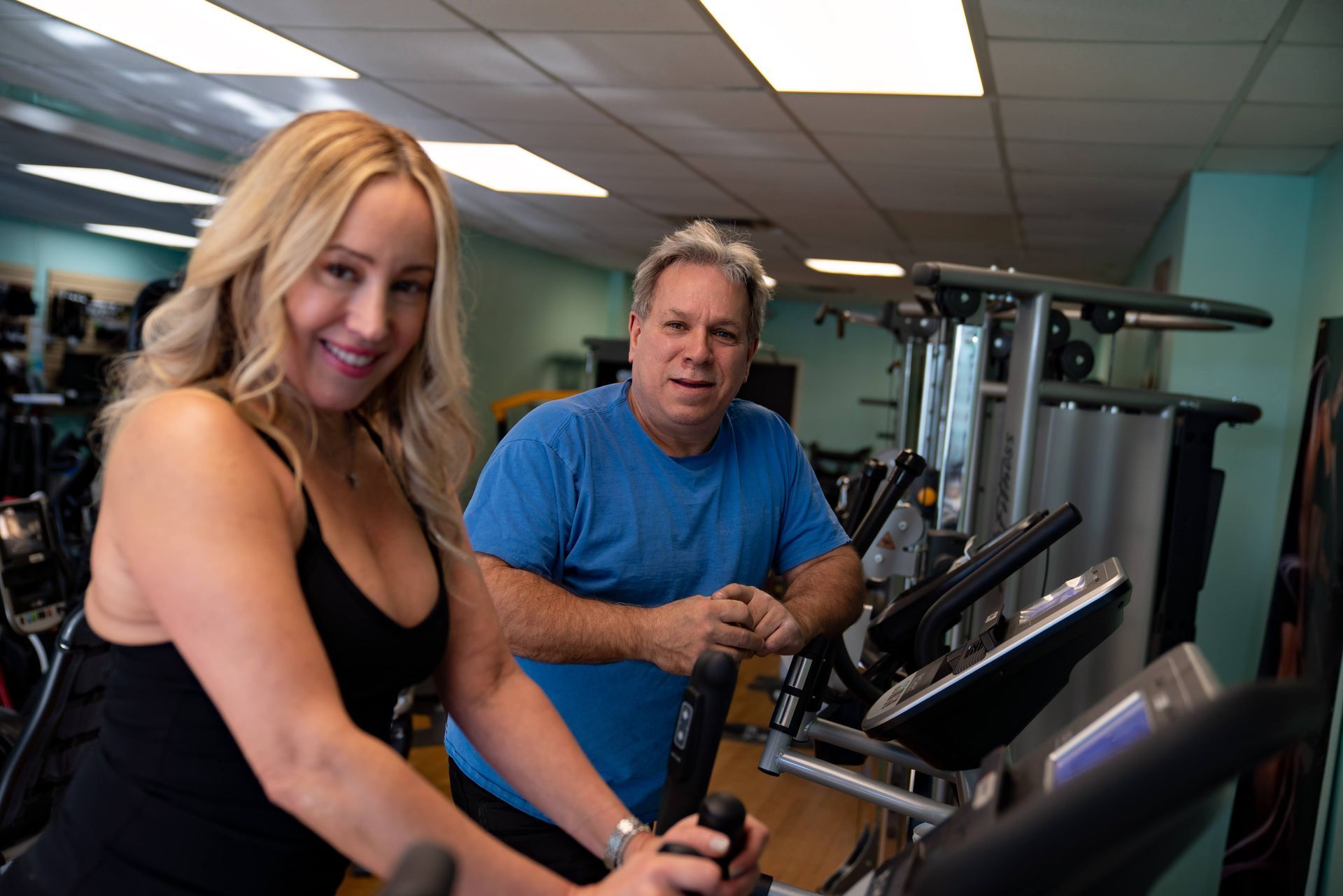 A blonde woman in a black tank top and a man in a blue t-shirt using elliptical machines at a brightly lit gym.