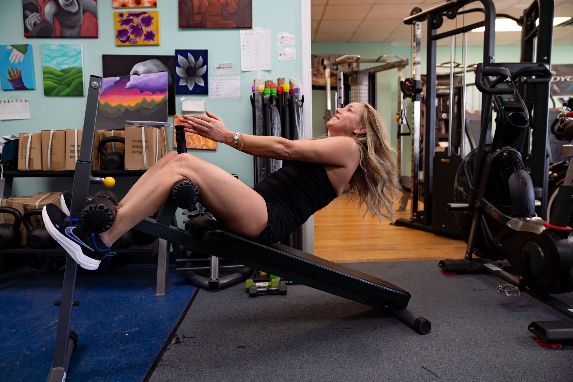 A person performs a sit-up exercise on an inclined bench inside a gym filled with fitness equipment and wall art.