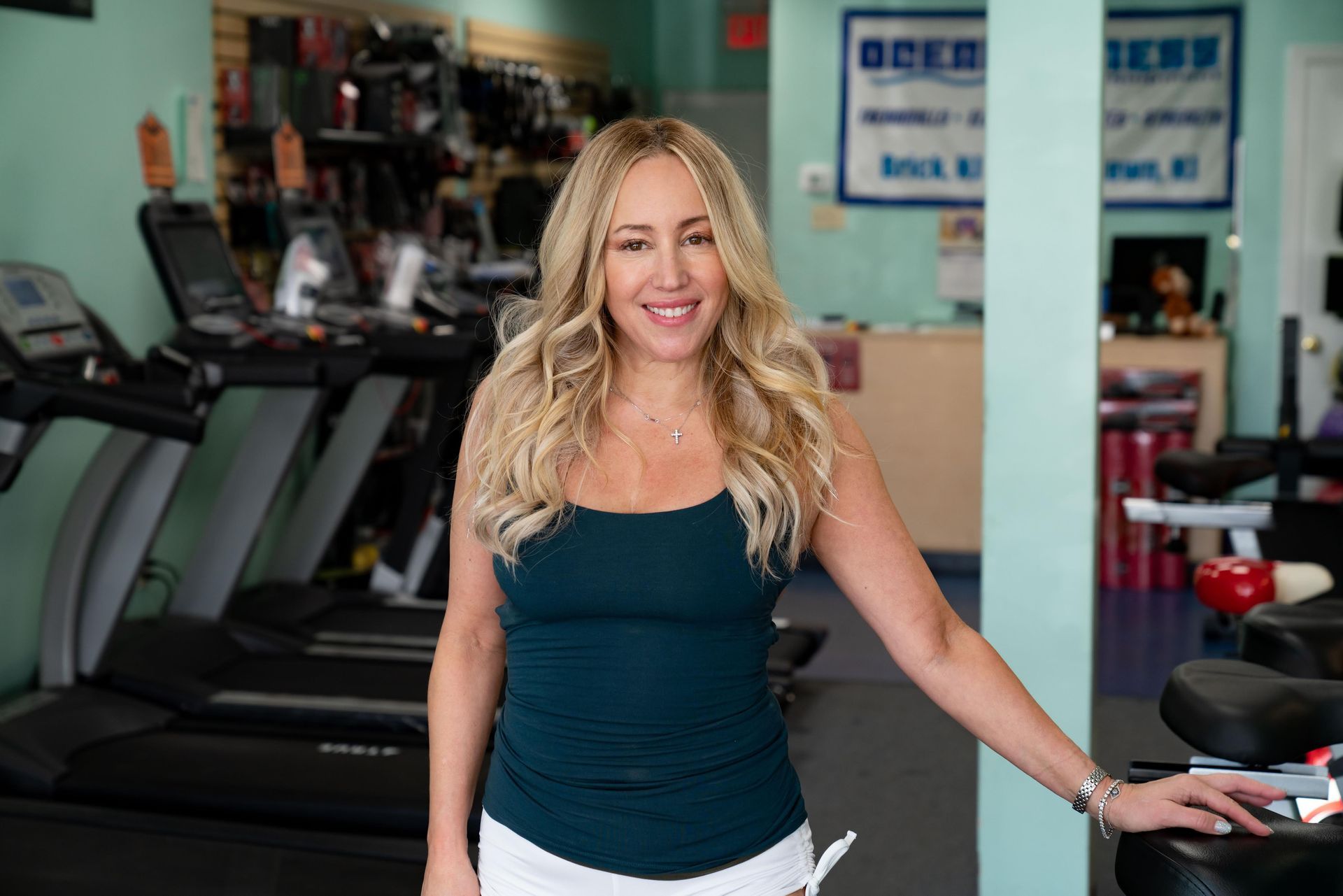 A smiling person in a dark tank top stands inside a gym, with treadmills visible in the background.