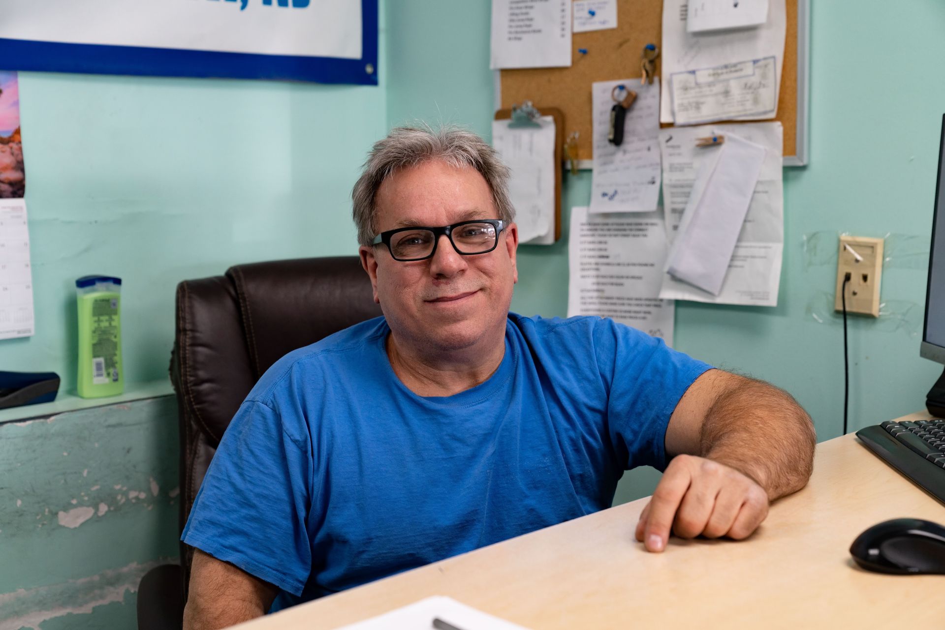 A person wearing glasses and a blue shirt sitting at a desk with a corkboard in the background.