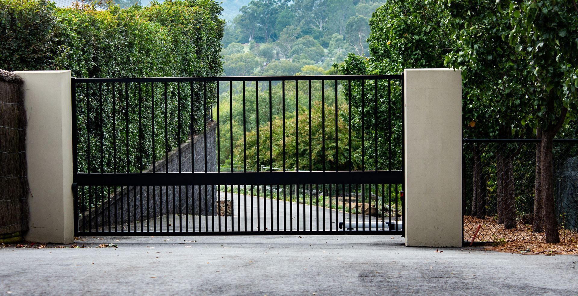 A black gate is open to a driveway with trees in the background.