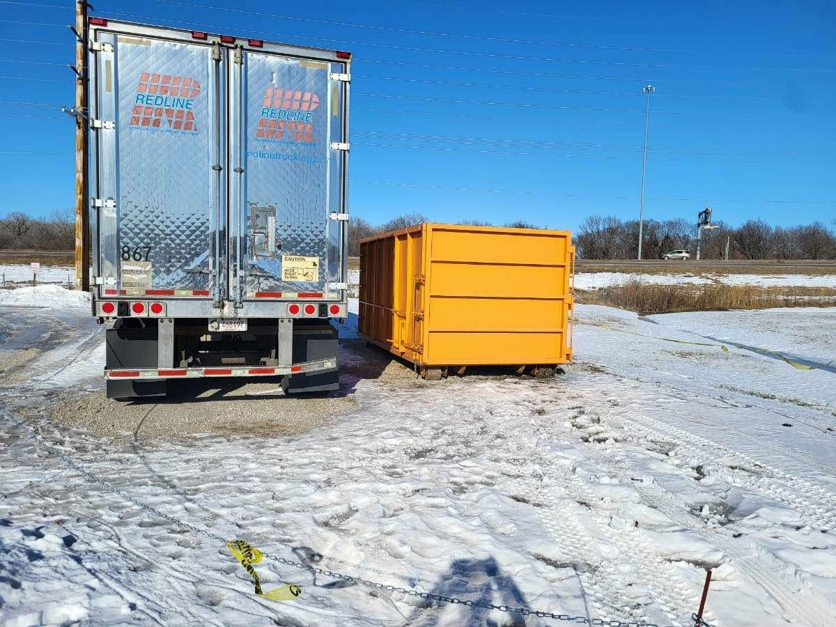 A shiny semi-trailer and a large yellow dumpster sitting side-by-side on a snow-covered, gravel-lined lot under a blue sky.