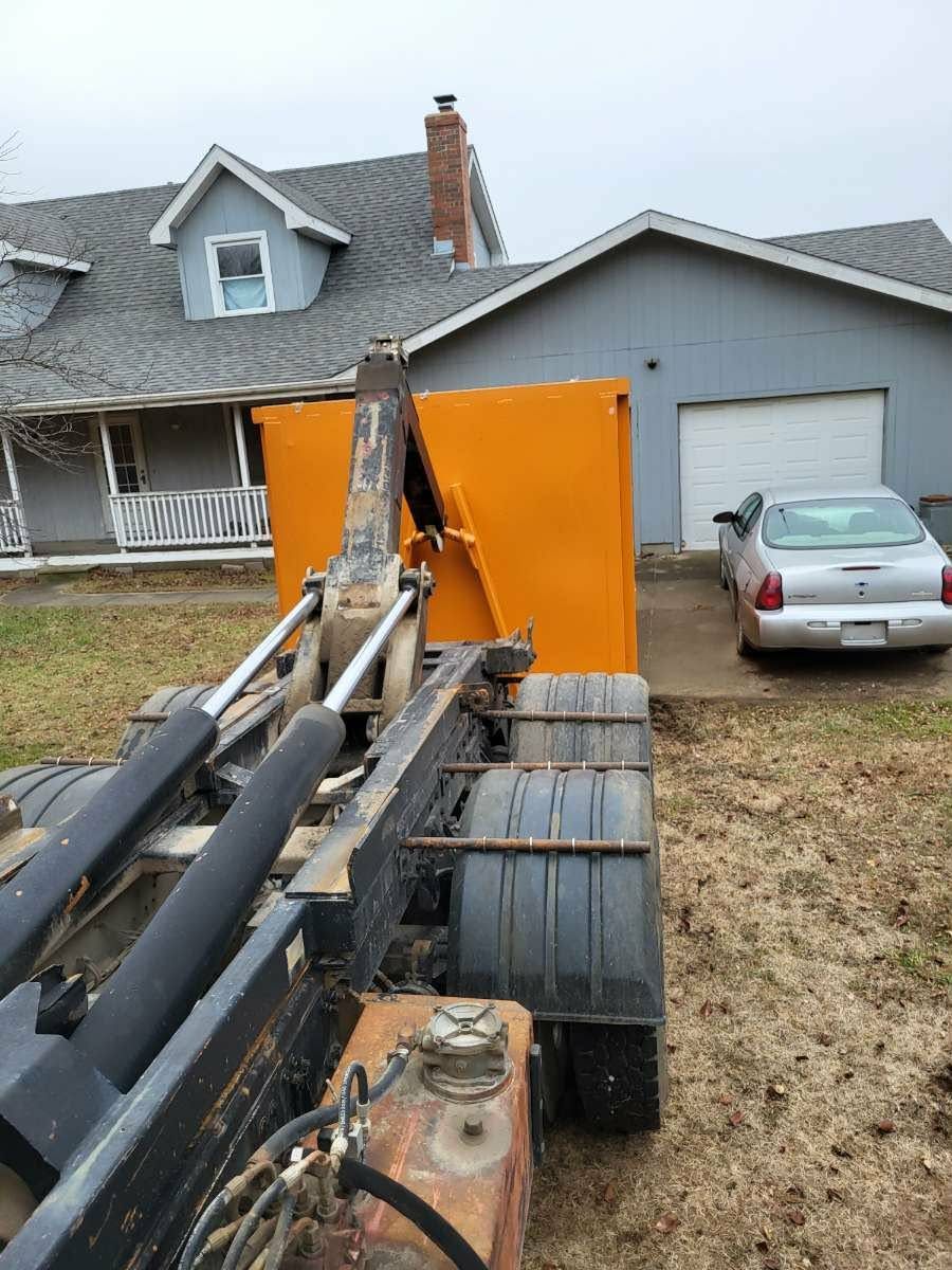 A roll-off dumpster truck backing up to place a bright orange dumpster in a residential driveway near a gray house.