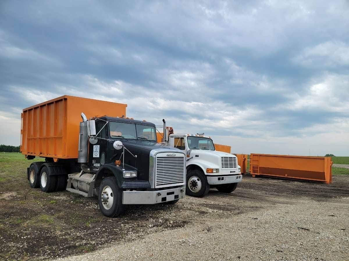 A black truck with an orange roll-off container and a white truck parked side-by-side on a gravel lot under a cloudy sky.