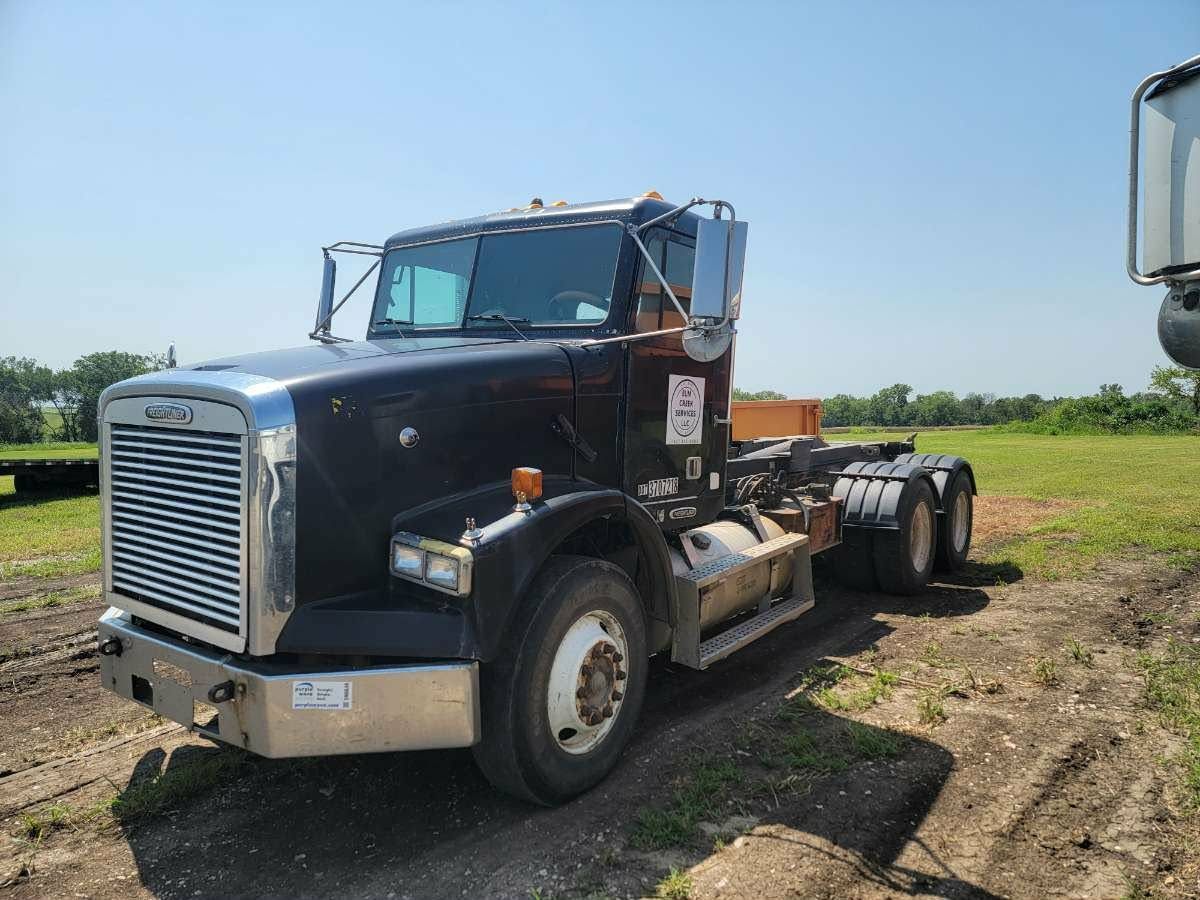 A black Freightliner semi-truck parked on a dirt lot in a rural, grassy field under a clear blue sky.