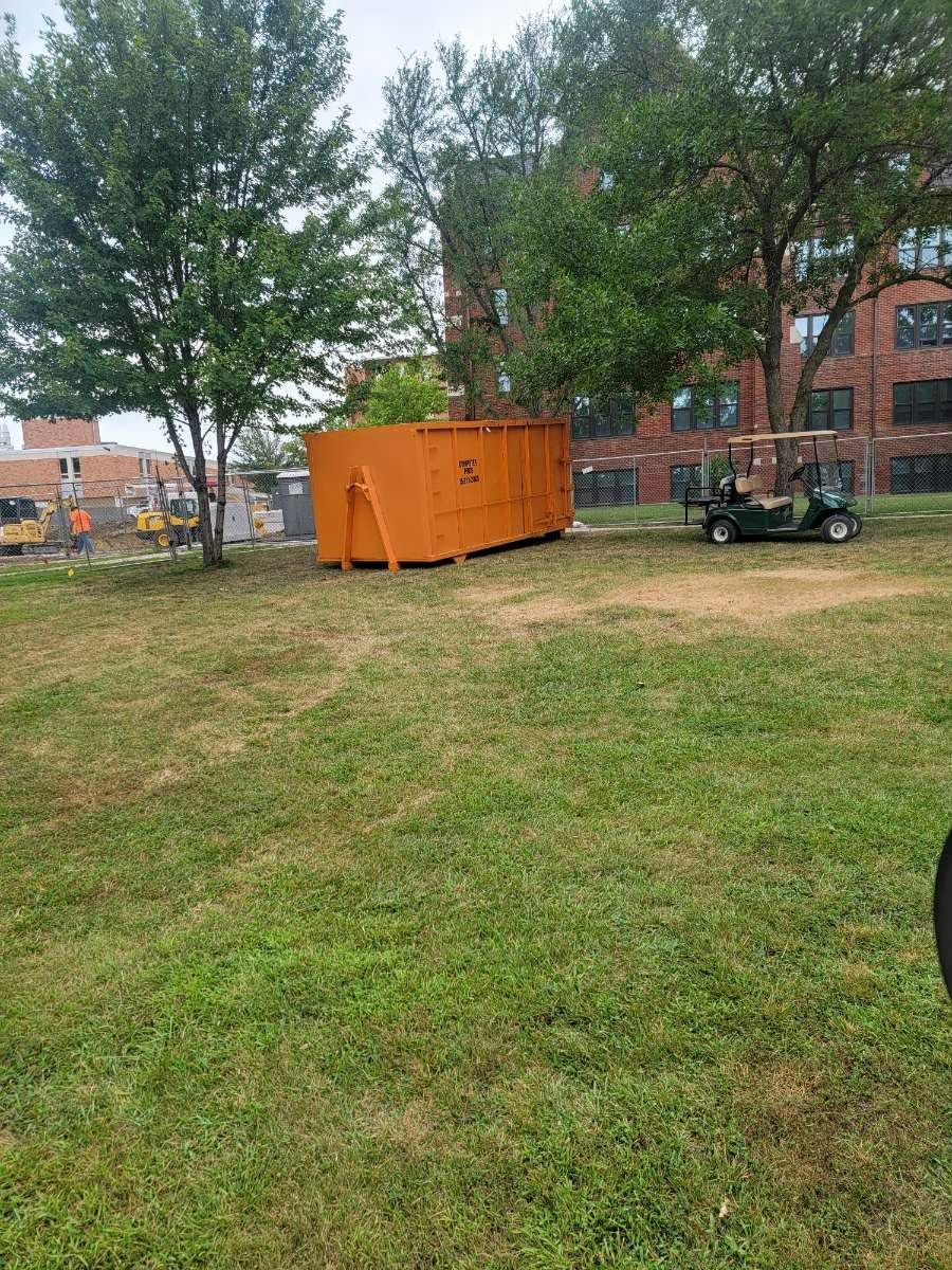 A bright orange roll-off dumpster sits in a grassy field near large trees and a multi-story brick building.