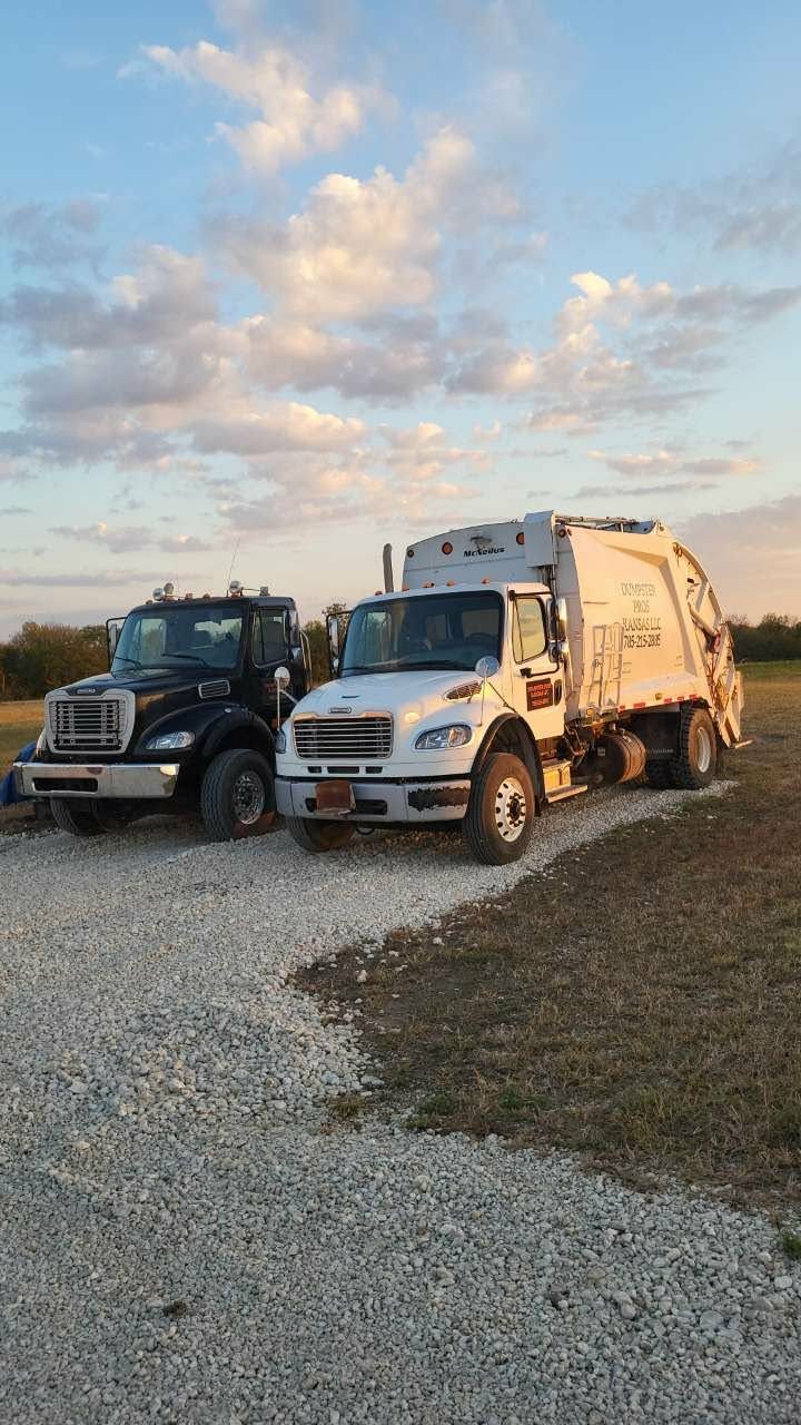 A black semi-truck and a white garbage truck parked side-by-side on a gravel lot at sunset.