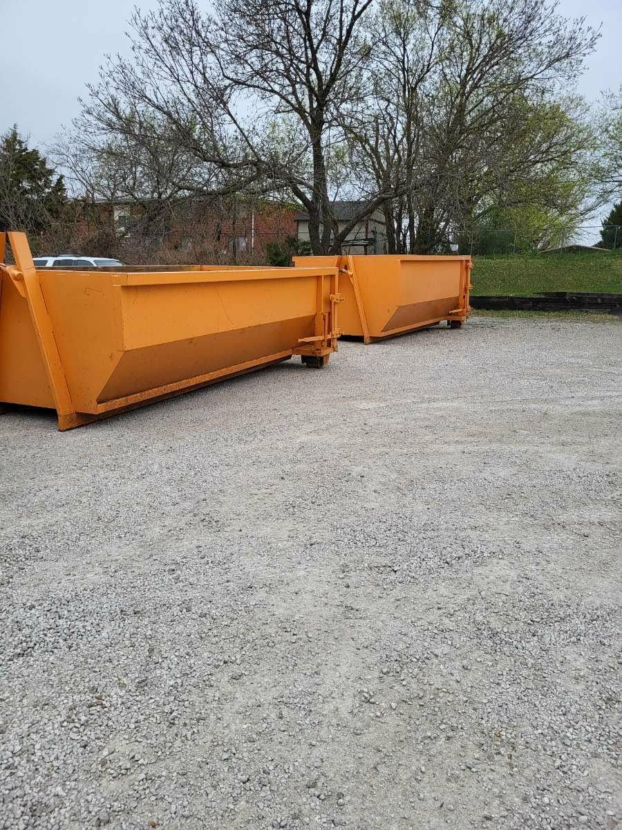 Two bright orange roll-off dumpsters sit empty on a gravel lot in front of trees and a brick building.