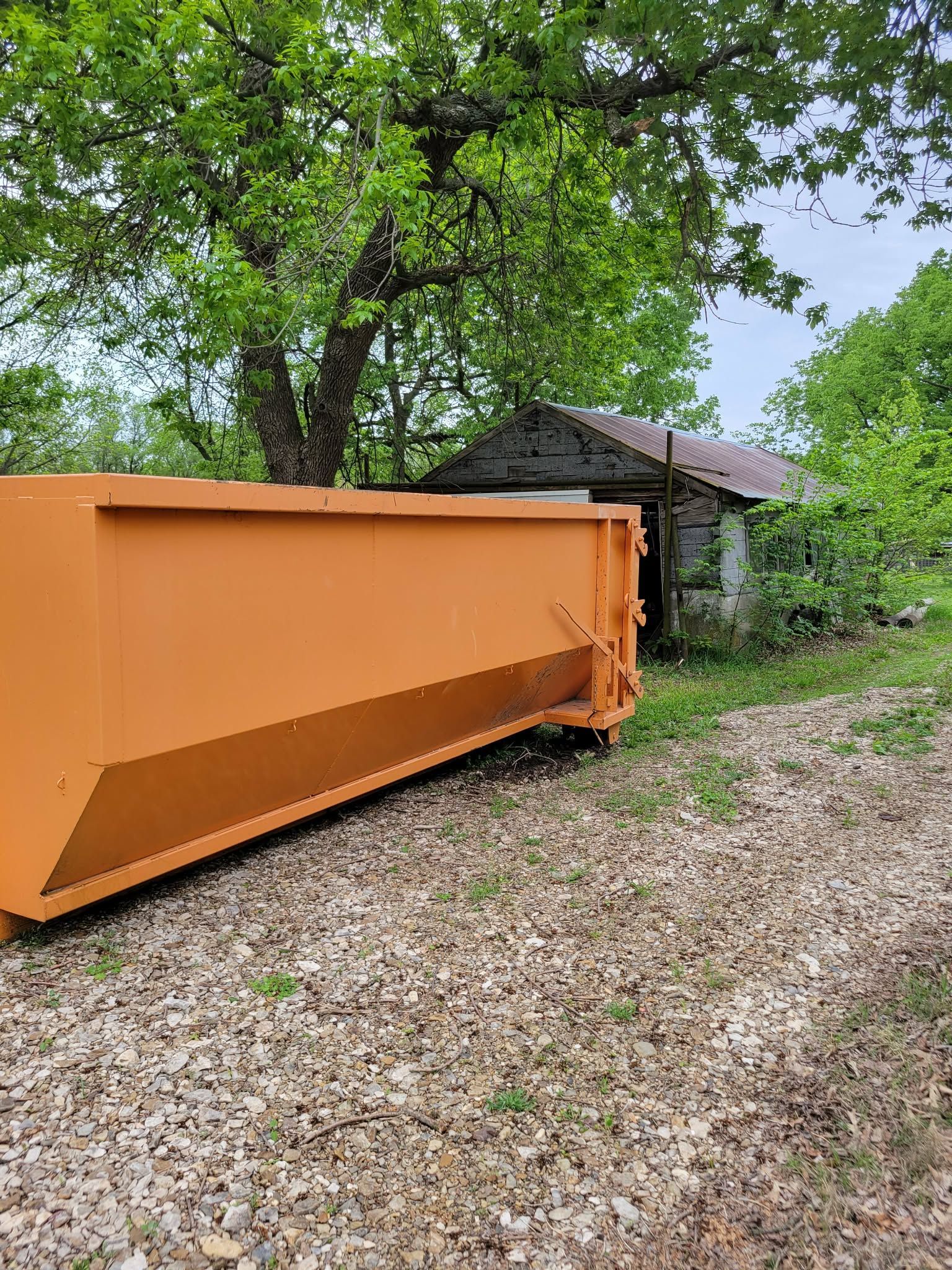 A large orange dumpster sits on a gravel path in front of an old, stone building surrounded by trees.
