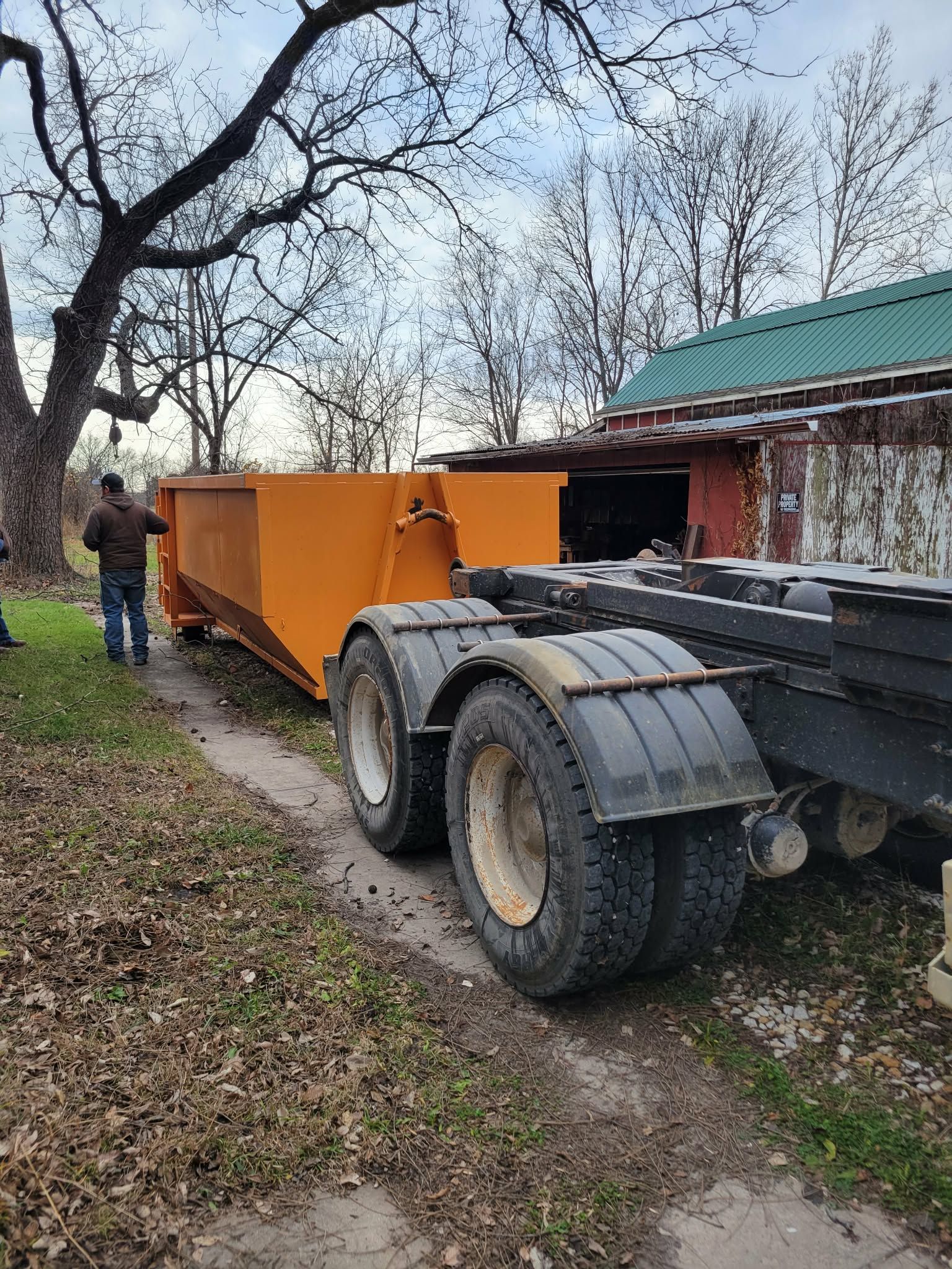 A person standing next to a large, bright orange roll-off dumpster being loaded onto a truck on a leaf-covered path.