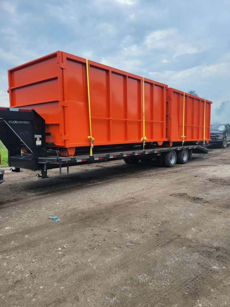 Two bright orange roll-off dumpsters secured with yellow straps on a flatbed trailer parked on a dirt lot.
