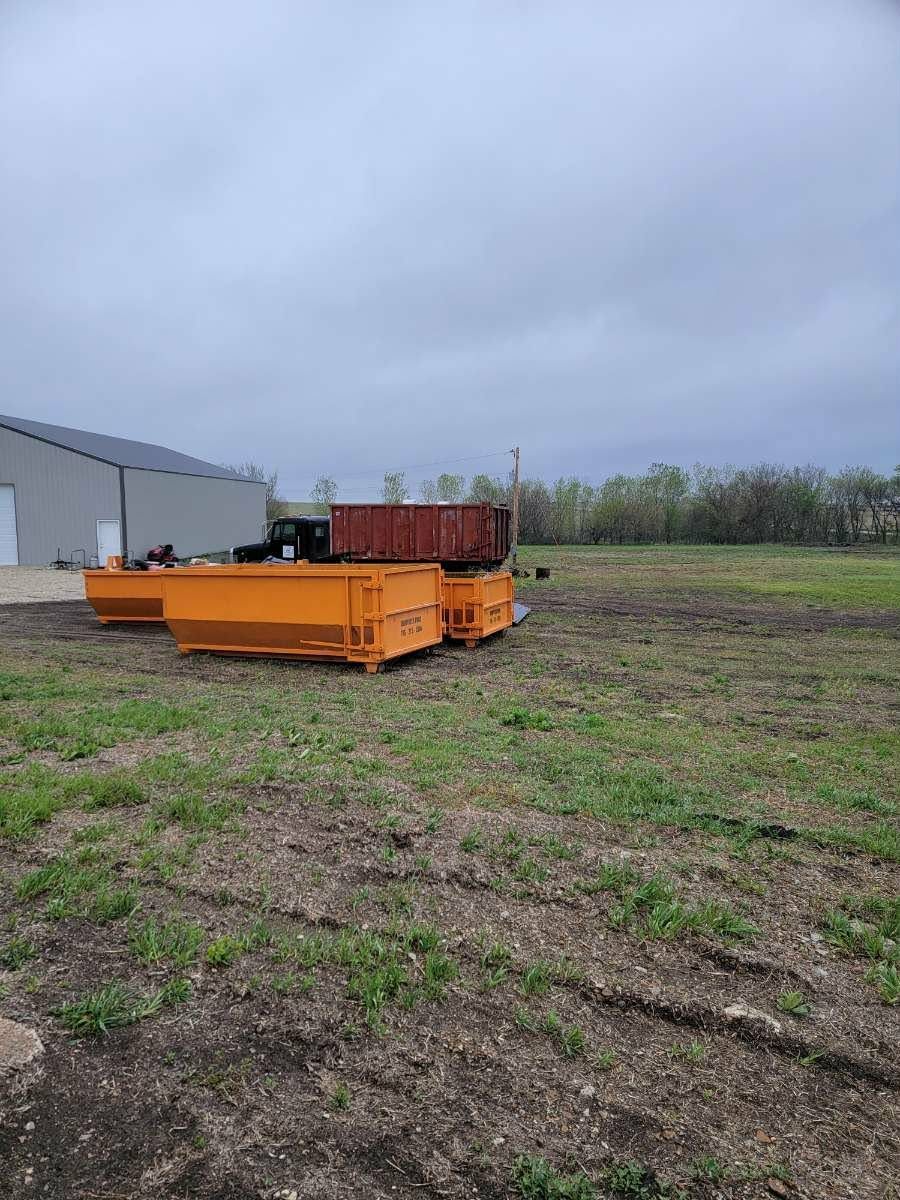 Bright orange dumpsters sit in a large, grassy field near a grey metal building under an overcast sky.